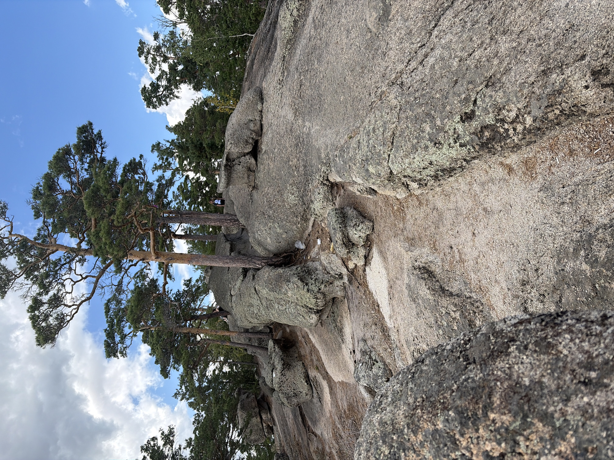 This image depicts a rocky landscape with scattered pine trees under a partially cloudy blue sky. In the mid-ground, a person wearing a dark top and light-colored shorts is visible sitting on a large boulder, facing away from the camera. Another person is standing further back on a rock formation, also facing away. The terrain is characterized by large granite boulders, with sandy patches and pine needles on the ground. The vegetation consists primarily of mature pine trees with visible bark texture and greenery. The scene appears to be an outdoor natural area, possibly a hiking or recreational spot within Зелёный Бор ауылдық округі, Kazakhstan.