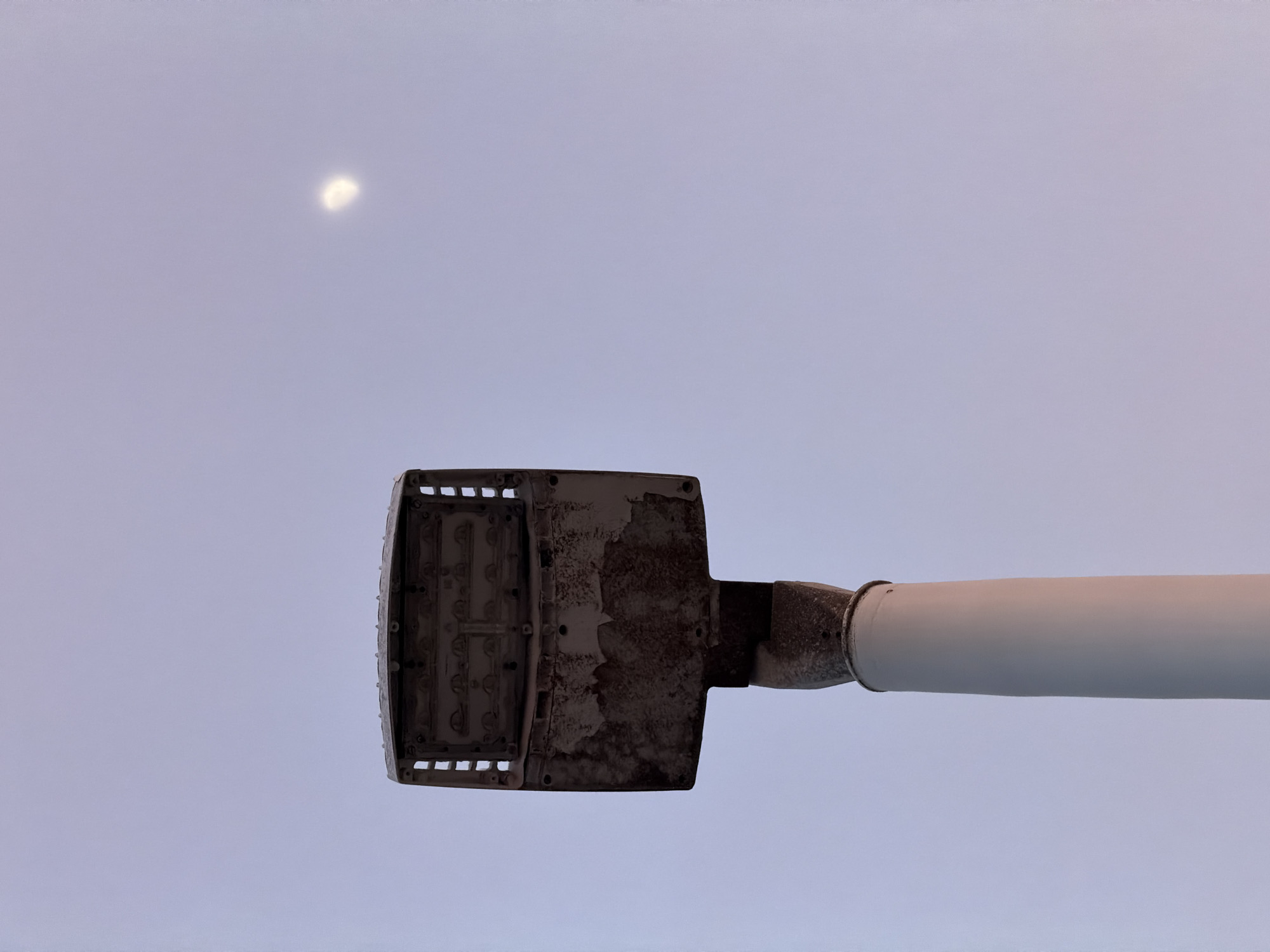 An upward shot of a street lamp post against a pale sky. The street lamp's housing is partially open, revealing what appear to be LED components. The housing is weathered and stained with rust. The pole of the street lamp is smooth and white, with a metal connector to the lamp housing that also appears weathered. A faint crescent moon is visible in the upper right quadrant of the sky. The scene is likely located in Miami Beach, United States of America, as indicated by the presence of a street lamp in a coastal urban environment.