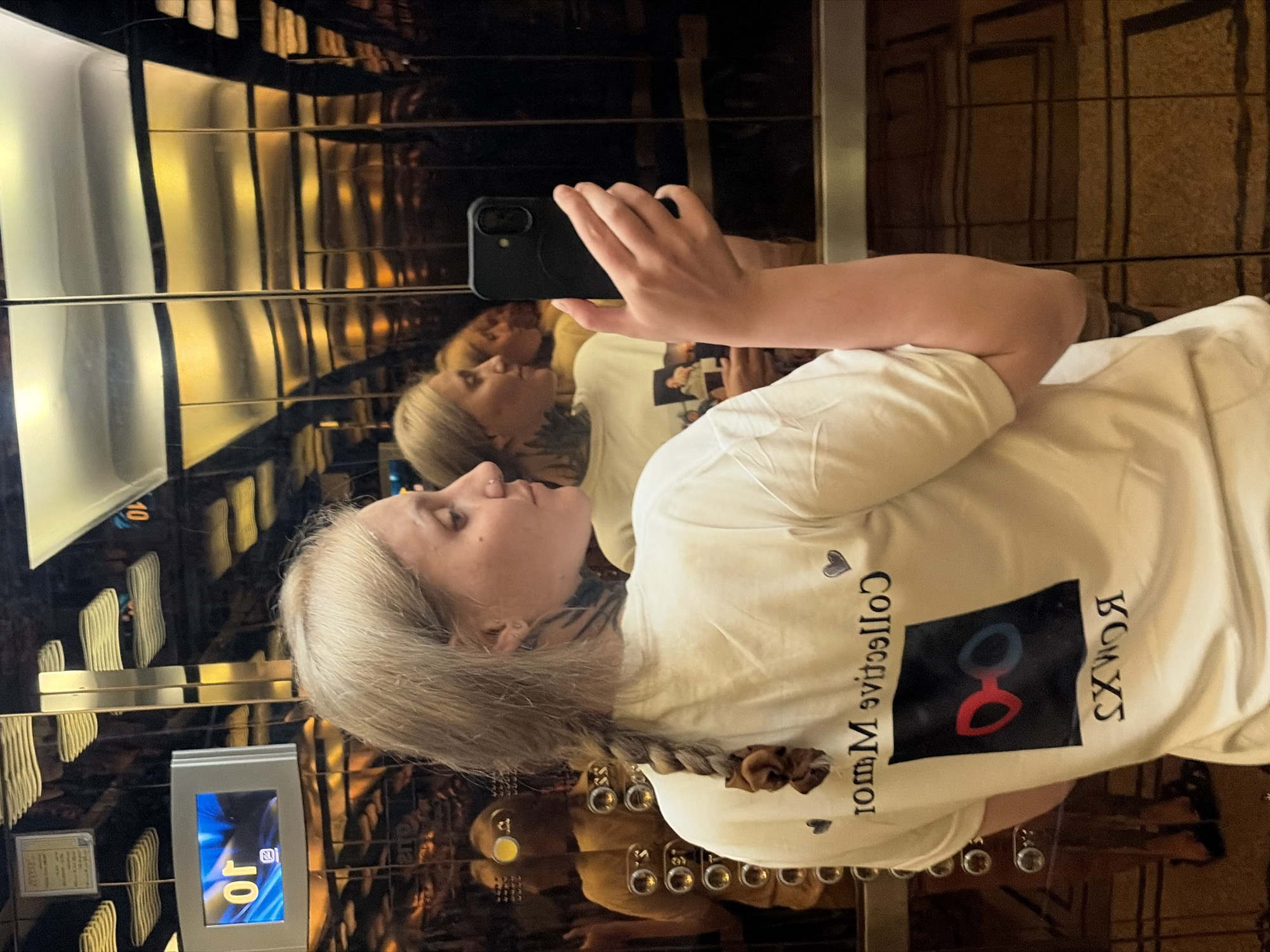 A woman with light blonde hair is taking a selfie in an elevator. She is wearing a white t-shirt with the text "Collective Mirror" and an image of glasses on it. The elevator has gold and black patterned walls. The floor display on the left shows the number 10. The elevator is located in Đà Nẵng, Vietnam.