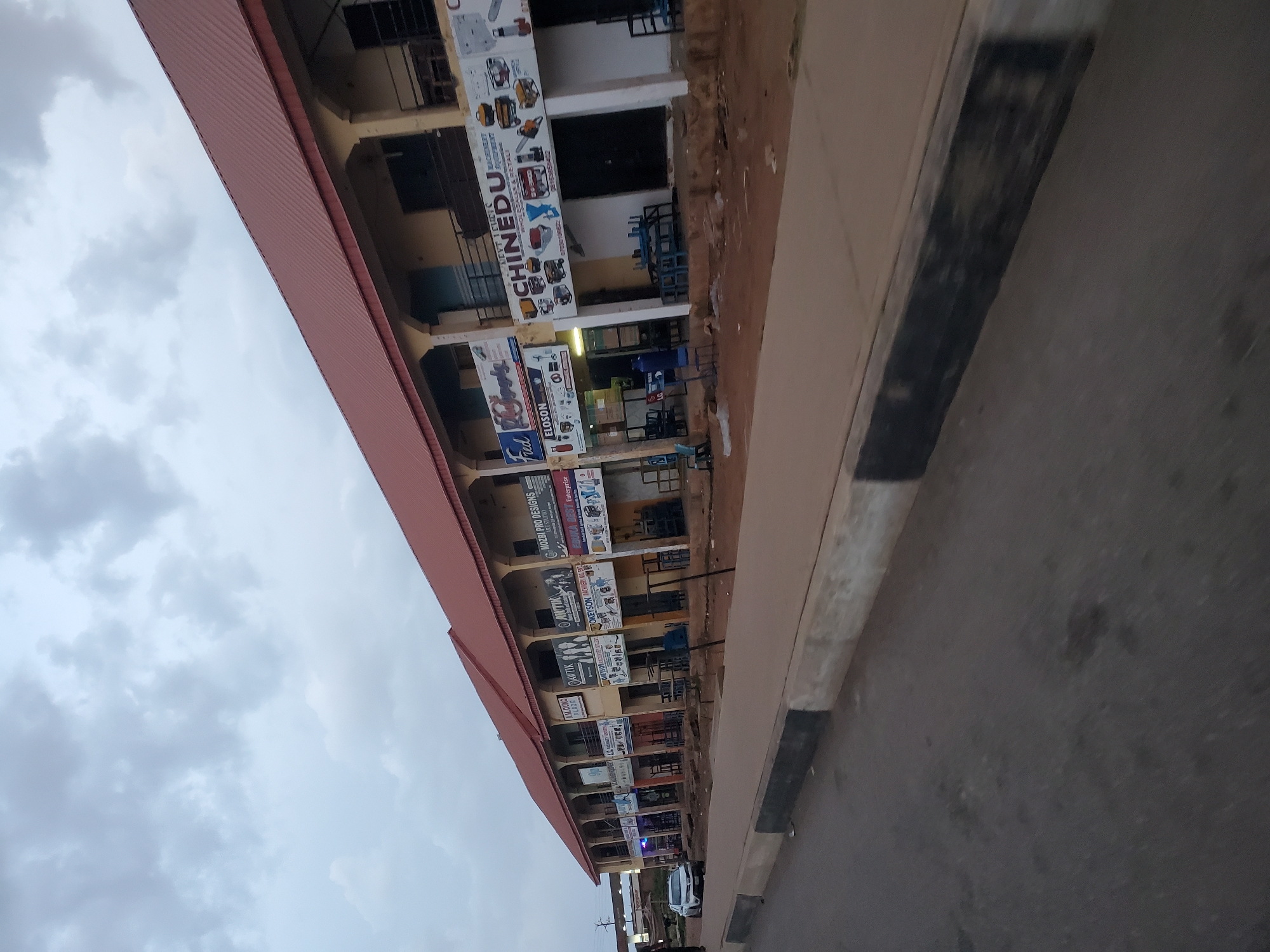 This image depicts a commercial strip in Gura Top, Nigeria, under an overcast sky. The buildings are low-rise with a continuous red tiled roof. Each unit appears to be a shop or business, with signage displayed prominently above their entrances. The facades are painted in a light yellow color. Many of the storefronts are open, revealing metal shelving and display units, suggesting a focus on retail. Some businesses are displaying images of machinery and equipment, indicating possible sales of industrial or agricultural tools. The ground in front of the shops is unpaved and dusty, with scattered debris. A paved sidewalk runs in front of the buildings, and beyond that, a paved road is visible, with a concrete curb separating the road from the sidewalk. A white car is parked on the side of the road. The overall impression is a functional commercial area, likely a local marketplace or business district.