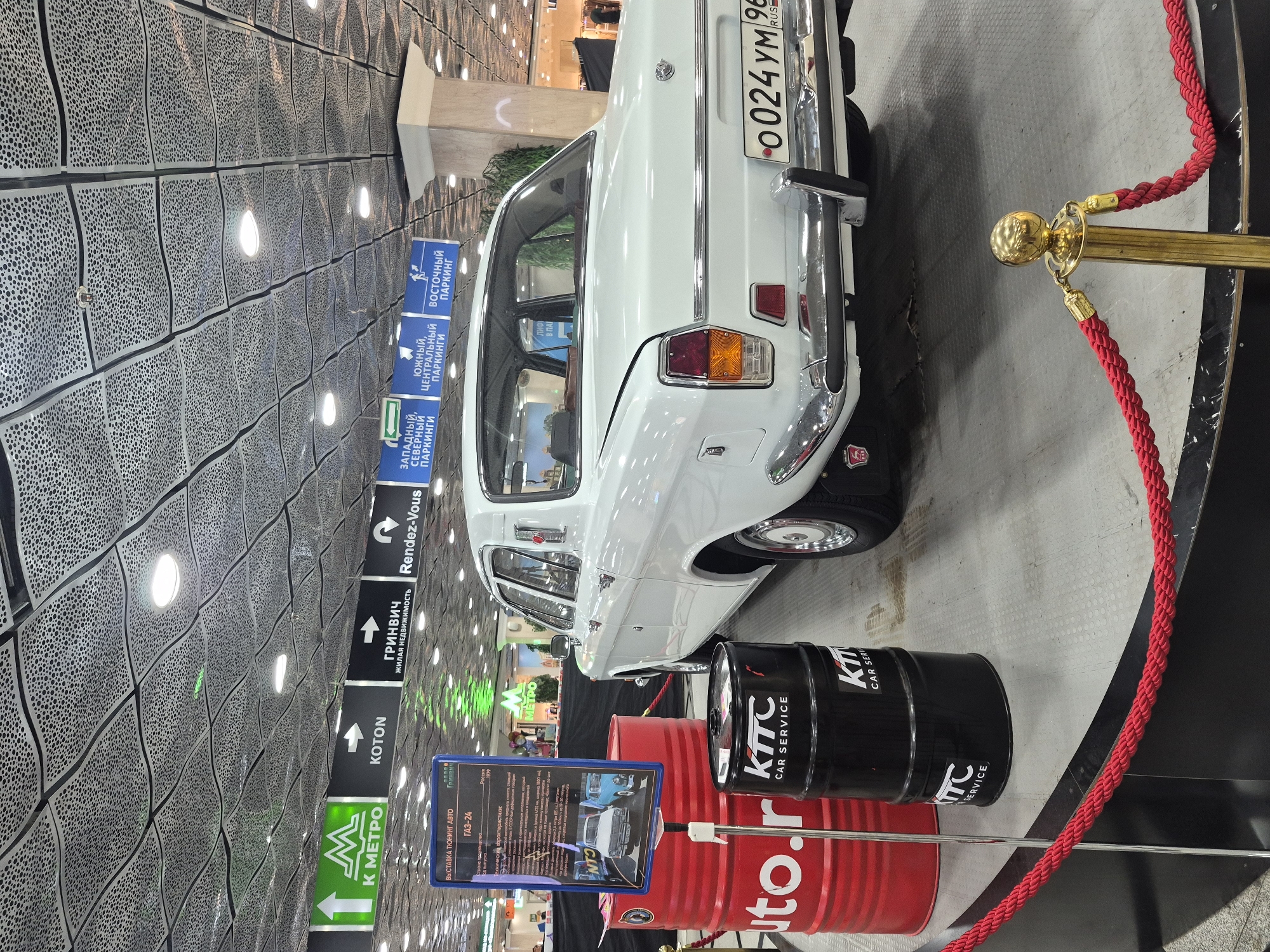 The image shows a white GAZ-24 sedan on display at an exhibition, likely in a shopping center or convention hall, in Yekaterinburg, Russia. The car is positioned on a raised platform, with red velvet ropes and gold stanchions cordoning off the display area. To the left of the car, there are two large red oil drums with "auto.r" printed on them and a black oil drum with "KIIC CAR SERVICE" branding. A blue information stand for the GAZ-24, displaying technical specifications and images, is also present. The background features a distinctive ceiling with a metallic perforated design and directional signs for various locations, including "K METRO," "KOTON," "GRINVICH," "Rendez-Vous," and parking areas labeled as "WESTERN, NORTHERN PARKING," "SOUTHERN, CENTRAL PARKING," and "EASTERN PARKING." The lighting is artificial, emanating from overhead fixtures. No people are clearly visible in the immediate vicinity of the car, though indistinct figures may be present in the further background. The overall scene suggests a public event or exhibition focused on vintage automobiles.