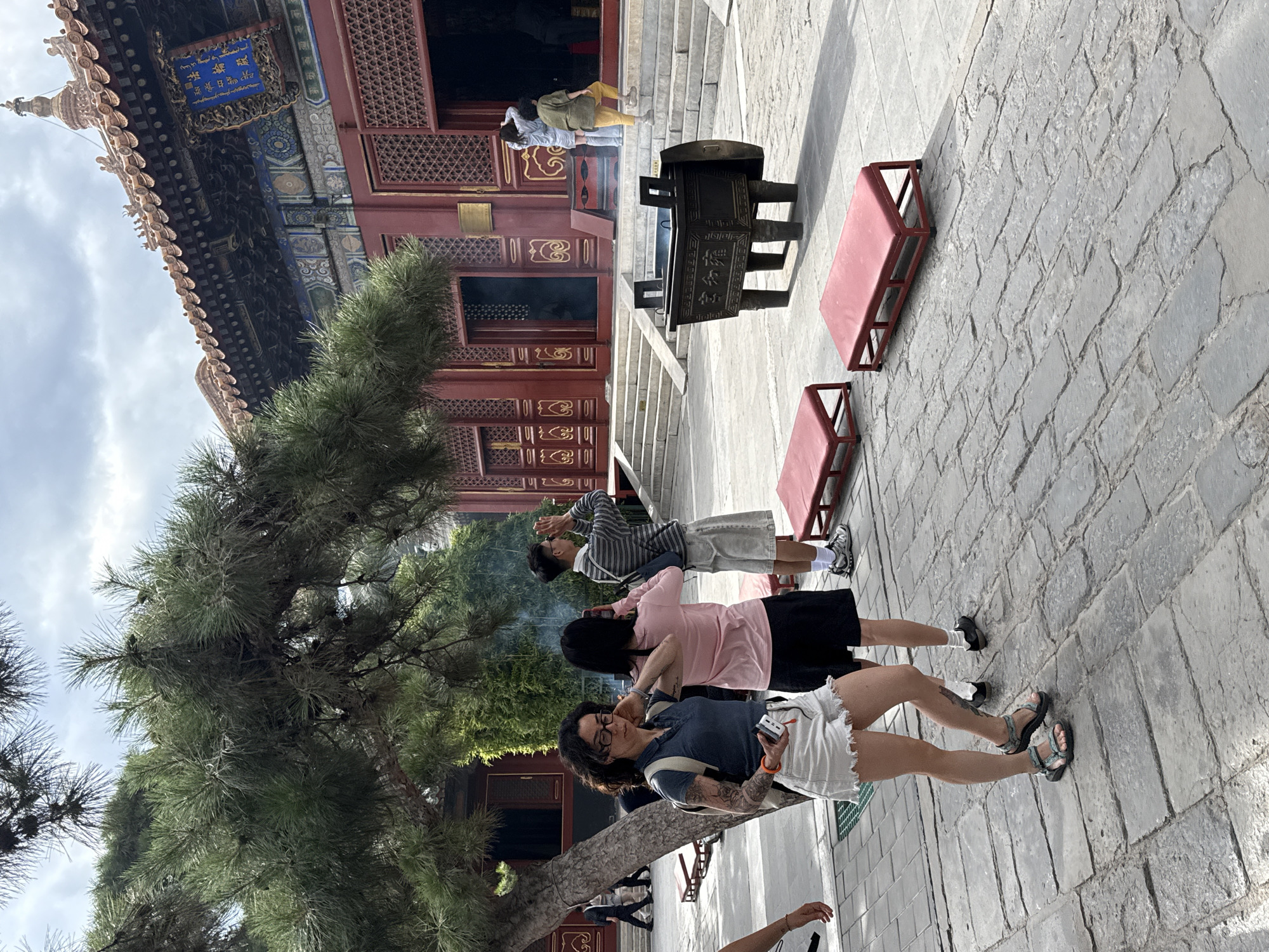 This image depicts a courtyard scene in the Dongcheng District, China, likely within a temple or palace complex, given the traditional architecture. A large pine tree with dense needles partially obscures the view of a red-painted building with ornate eaves and a blue plaque. Several people are present in the courtyard, some appearing to be tourists and others engaged in prayer or observation.
In the foreground, a woman with dark curly hair and glasses stands facing the camera, wearing a blue polo shirt, white denim shorts, and sandals. She has tattoos on her left arm and is carrying a backpack. To her right, another woman in a pink shirt and black skirt is seen from behind, holding a phone. Further back, a man in a striped shirt and shorts stands with his hands clasped in front of his face, seemingly in prayer.
In the middle ground, a large, ornate bronze censer sits on the paved courtyard. Red benches are placed around the courtyard. In the background, steps lead up to the entrance of the red building, where more individuals are visible, some entering the structure. Smoke appears to be rising from inside the building, possibly from incense. The ground is paved with large, irregular stone slabs. The sky is overcast.