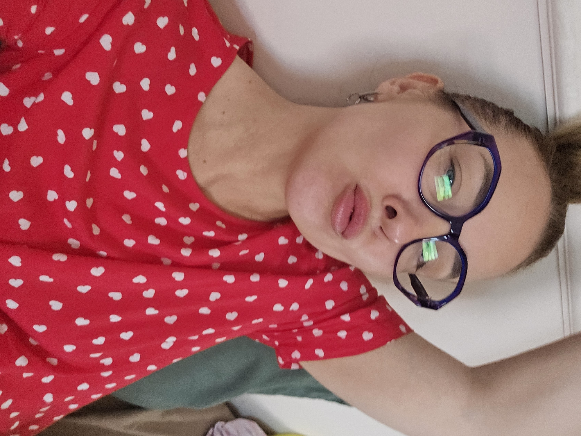 A woman is lying down in what appears to be an indoor setting. She is wearing a red top with white heart-shaped patterns and blue-rimmed glasses. Her hair is pulled back in a bun. The background is a plain white wall. There is no visual indication of the location being Sochi, Russia or any other specific city.