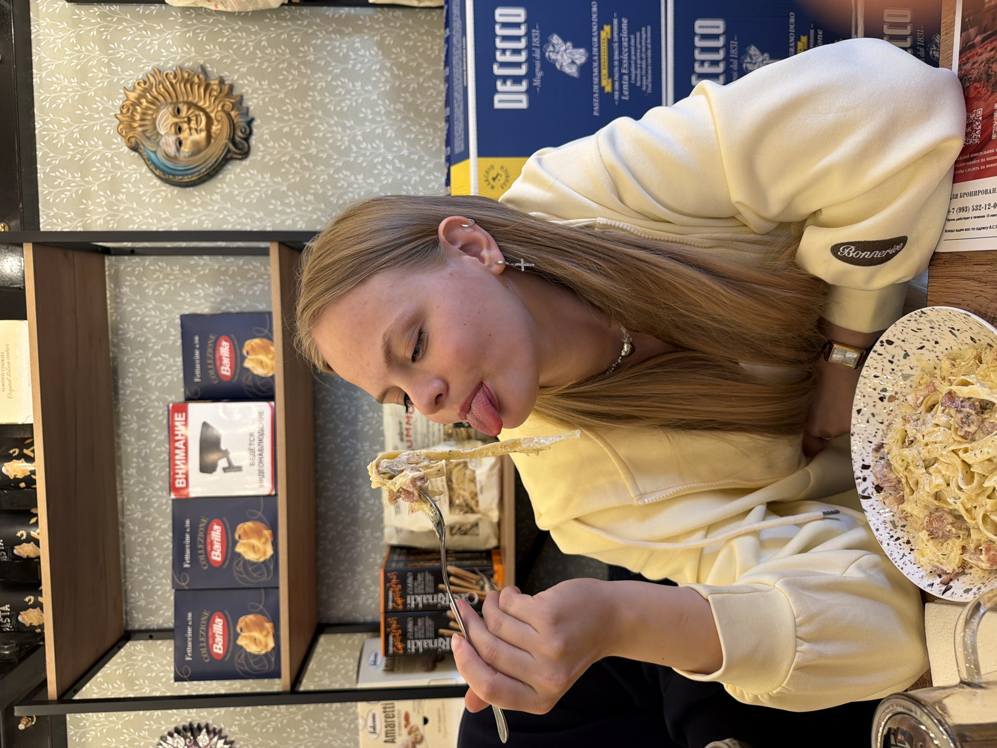 The image shows a young woman in an indoor setting, likely an Italian-themed restaurant or cafe. She has long, straight, light brown hair and is wearing a light yellow hooded sweatshirt with "Bonnertee" embroidered on the sleeve. She is captured mid-meal, holding a fork piled with pasta, sauce, and what appears to be bacon or ham, and playfully sticking her tongue out. Her left ear has multiple piercings, including a cross earring. A watch is visible on her right wrist.
The background features shelves stocked with Italian food products, reinforcing the setting. Prominent brands include large blue boxes of "DE CECCO" pasta, labeled "PASTA DI SEMOLA DI GRANO DURO" and "Lenta Essiccazione," along with smaller blue boxes of "Barilla Collezione Fettuccine." Other products like "Rinaldi" breadsticks and "Amaretti" almond cookies by "Jaloons" are also visible. The walls are covered in a light green wallpaper with a white leaf pattern, and a golden sun-face decorative piece is mounted above the shelves.
A plate of pasta, similar to what the woman is eating, rests on the table next to her. Several signs with Russian text are visible, including "ВНИМАНИЕ" (Attention) and "ВЕДЕТСЯ ВИДЕОНАБЛЮДЕНИЕ" (CCTV in progress) on a red-bordered sign. Another sign displays a Russian phone number for reservations, "+7 (993) 532-12-0," and invites customers to "ПОДПИСЫВАЙСЯ на наши" (Subscribe to our...). The presence of Italian brand names and Russian text confirms the location as an Italian-themed eatery in Russia, specifically Izhevsk as per the provided context. The activity portrays a casual and playful moment during a meal.