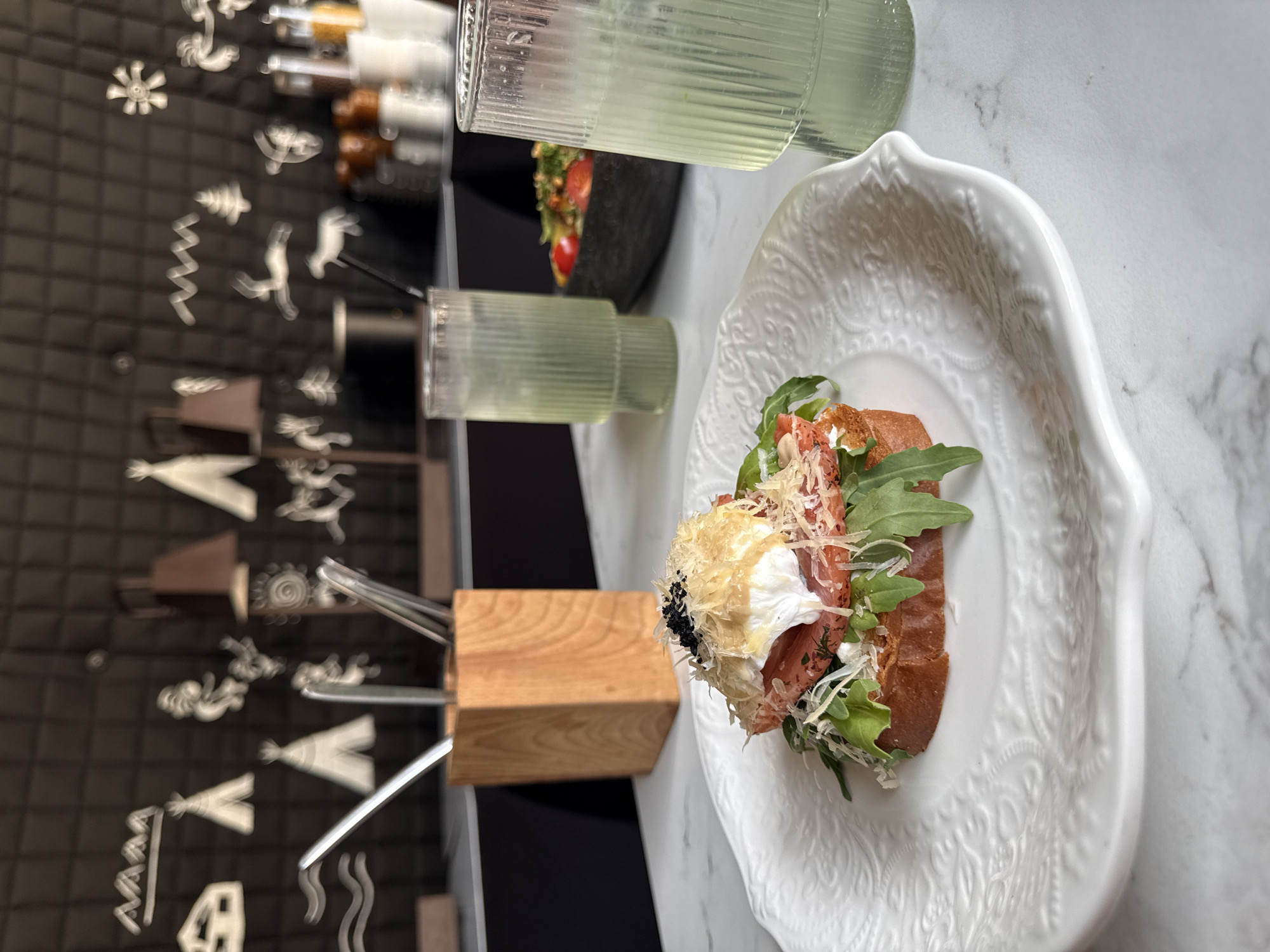 This is a medium shot photograph taken indoors at a restaurant in Ust'-Muny, Russia. The focal point is a slice of toast topped with a poached egg, salmon, and grated cheese, served on a white, ornate plate. In the background, two tall glasses filled with a pale green beverage are visible, one with a straw. A bowl of salad with tomatoes and greens is also present. The restaurant's interior features a dark wall adorned with white, stylized drawings of tents, animals, and abstract shapes, suggesting a theme. A wooden holder containing utensils sits on the table. The table itself is marble-patterned. The lighting appears to be artificial, creating a slightly dim ambiance.
