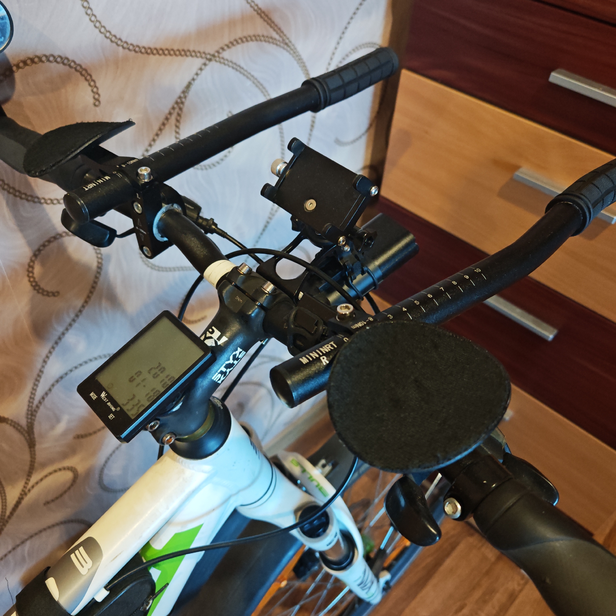 The image depicts a close-up view of a bicycle's handlebars and front frame. The bike is mostly white with green accents. The handlebars are black and have various attachments including a phone mount, a cycling computer displaying information, and what appear to be aero bars with pads. The brand names "WEST BIKING," "MININRT," and "STYX" are visible on the equipment. The background consists of patterned fabric and a wooden dresser with metal handles. There is no information provided in the image to indicate that this picture was taken in Rostov, Russia.
