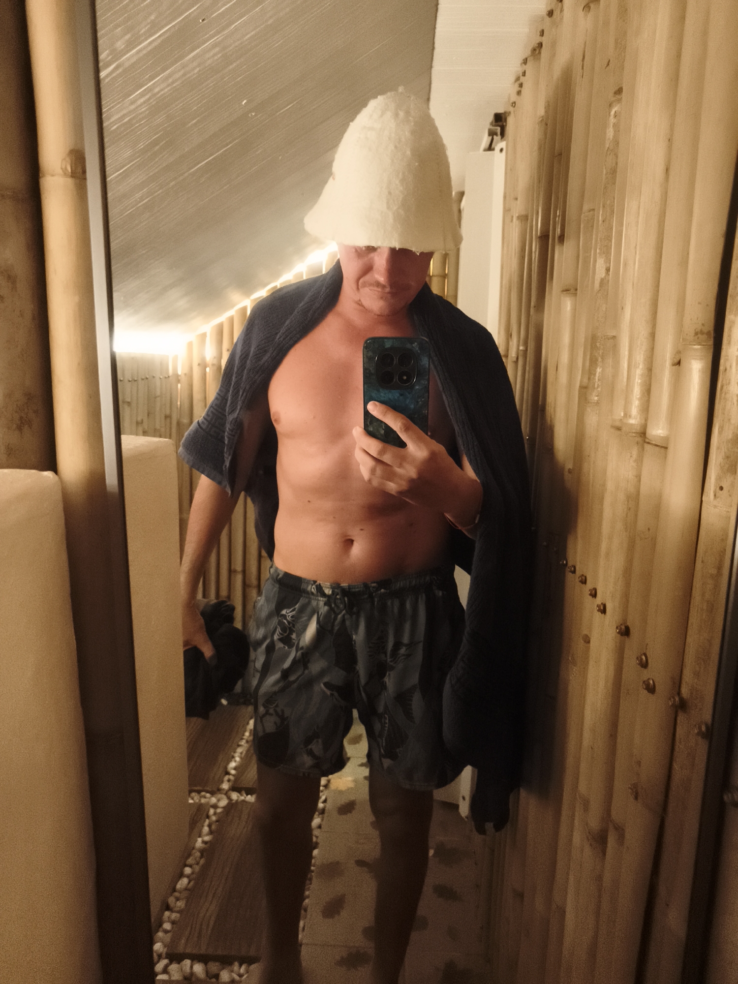 A man stands in a room with bamboo walls, holding a phone and taking a selfie in a mirror. He is shirtless, wearing patterned shorts, and has a towel draped over his shoulders and a white felt sauna hat on his head. The floor is made of wood planks with pebbles. The room appears to be a sauna. The location is บ้านบางรักษ์, Thailand.