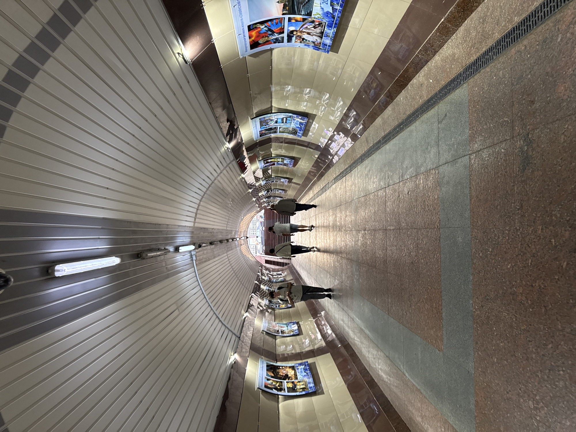 The image depicts a long, arched pedestrian tunnel with a metallic, ribbed ceiling. Fluorescent lights are mounted along the ceiling. The walls of the tunnel are tiled in alternating bands of cream and dark red, with posters displayed at intervals on both sides. The floor is paved with large, speckled red and grey tiles, with a drainage grate running along one side of the tunnel. Four individuals are walking away from the viewer down the tunnel. The furthest person is ascending stairs at the end of the tunnel, which opens to what appears to be an outdoor scene with buildings and cars visible. The overall impression is one of an urban underpass or subway station.
