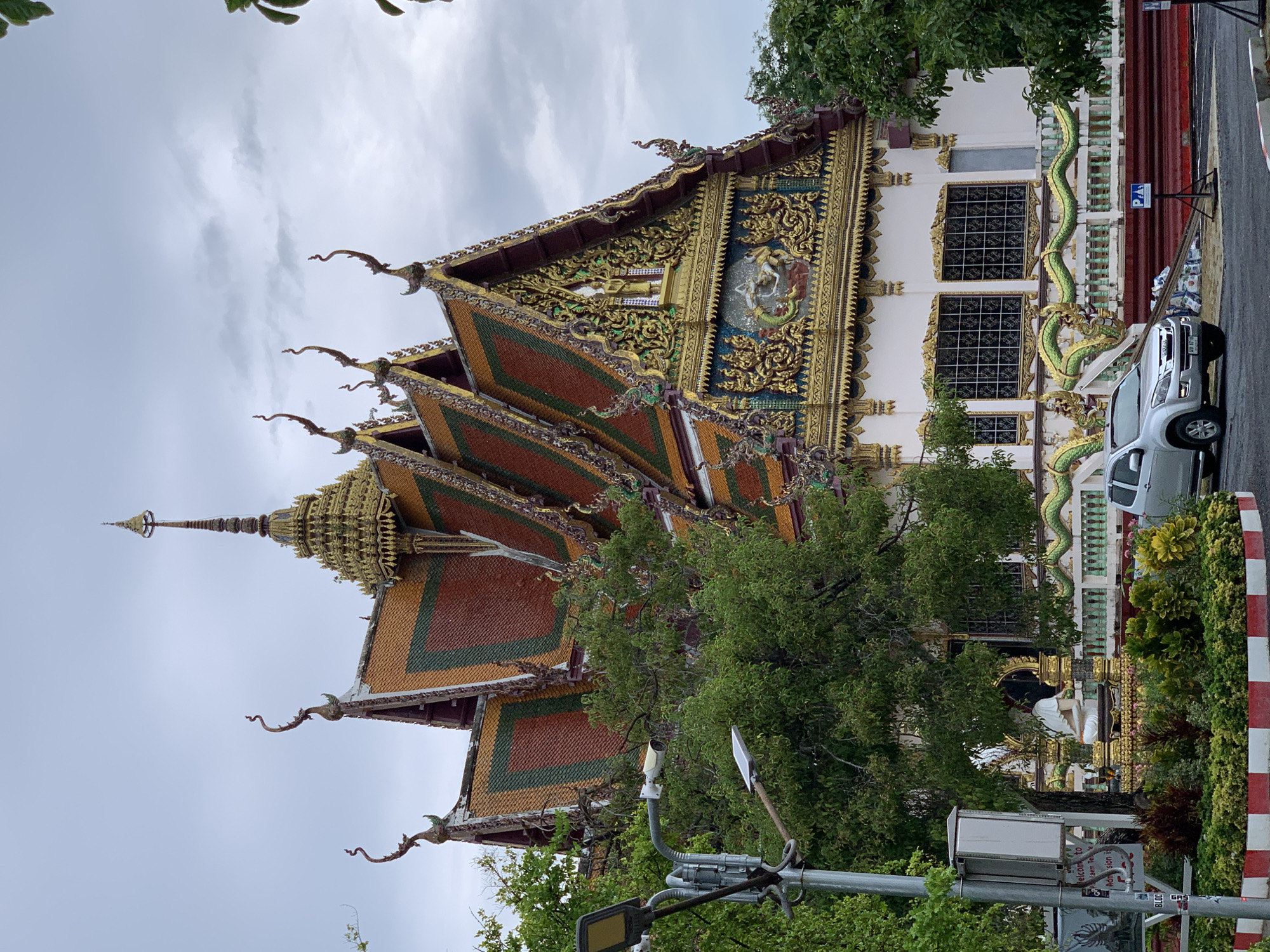 The image displays a grand and ornately decorated temple building, likely a Wat, situated in Baan Plai Laem, Thailand. The sky is overcast, suggesting a cloudy day.
The temple features a multi-tiered roof with vibrant, colorful tiles in shades of red, orange, green, and gold, intricately trimmed with golden ornamentation. Elaborate, upward-curving finials (chofahs) adorn the roof edges. A golden spire or chedi rises prominently from one of the temple's structures. The main gable end is a focal point, covered in rich golden stucco work, green and blue patterns, and a central relief sculpture depicting mythical or religious figures.
The temple's white facade is punctuated by large rectangular windows fitted with decorative grilles. A long, undulating green naga balustrade serves as a prominent architectural element along the front.
In the foreground and to the right, a paved road or driveway is visible, bordered by a red and white striped curb. A silver pickup truck, an Isuzu D-Max, is parked beside the road. Green trees and bushes are scattered throughout the scene, partially obscuring portions of the temple and the street. Street infrastructure includes a street light pole equipped with a security camera.
Visible text includes a blue parking sign with a white "P" symbol. A white sign on a pole, partially obscured, reads "Welcome to Baan Plai Laem Temple" and "Admission". A smaller sign below it appears to say "BLOCK GUS". The license plate on the pickup truck is in Thai script but is not clearly legible.