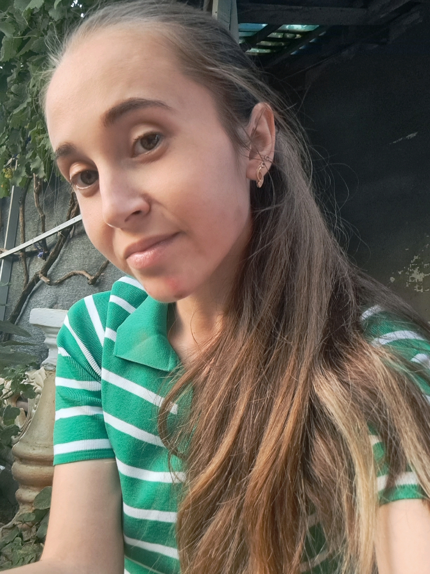 A young woman with long, brown hair, wearing a green and white striped shirt, is posing for a selfie outdoors. She has a slight smile and is looking directly at the camera. The background shows some greenery and a dark structure, possibly a wall or roof. The image appears to be taken in a residential area.