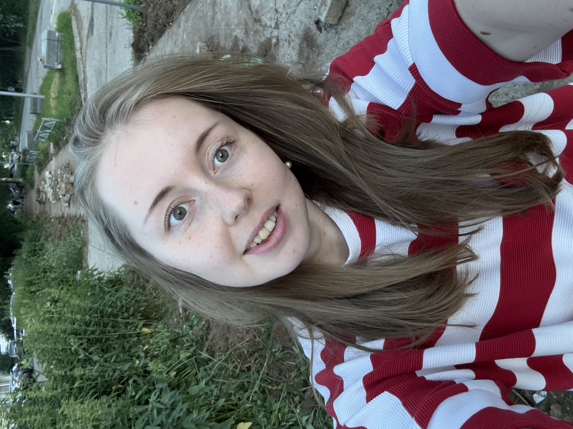 An exterior, eye-level, medium shot shows a young Caucasian woman with freckles and long, light brown hair. She is wearing a red and white horizontally striped long-sleeved shirt. She is smiling with her teeth showing, her head tilted to her right. Her left eye is looking directly at the camera. A single pearl earring is visible in her left ear. The background is blurred and appears to be an outdoor urban environment. A sidewalk is visible in the foreground, with some scattered rocks and debris. To the left of the sidewalk is an area of overgrown green plants. In the distance, a street with cars and people walking is visible, along with some construction materials. The lighting suggests it is daytime.