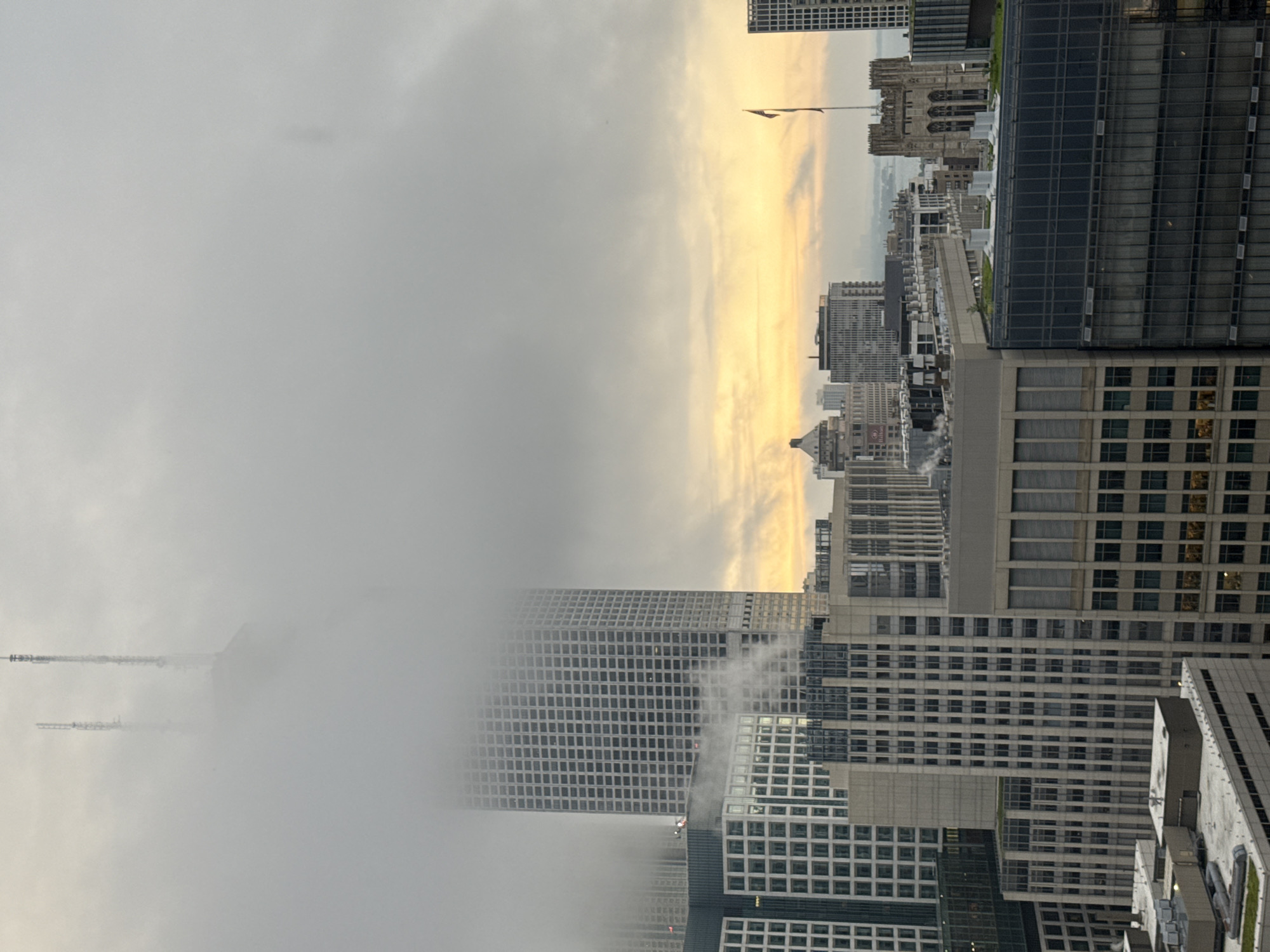 The image shows a view of the Chicago skyline shrouded in fog and clouds. The upper portions of several tall buildings are obscured by the fog, with only the tops of antennas visible on one skyscraper. The sky is a muted gray, transitioning to a warmer, hazy yellow and orange near the horizon, suggesting either sunrise or sunset. Several buildings are visible, including a large, grid-like skyscraper in the center-left, and a gothic-style building with a flag on its right. The foreground consists of various office buildings and rooftop structures, some with visible ventilation systems. No people are discernibly present in the image. The overall impression is of a dense, atmospheric urban environment.