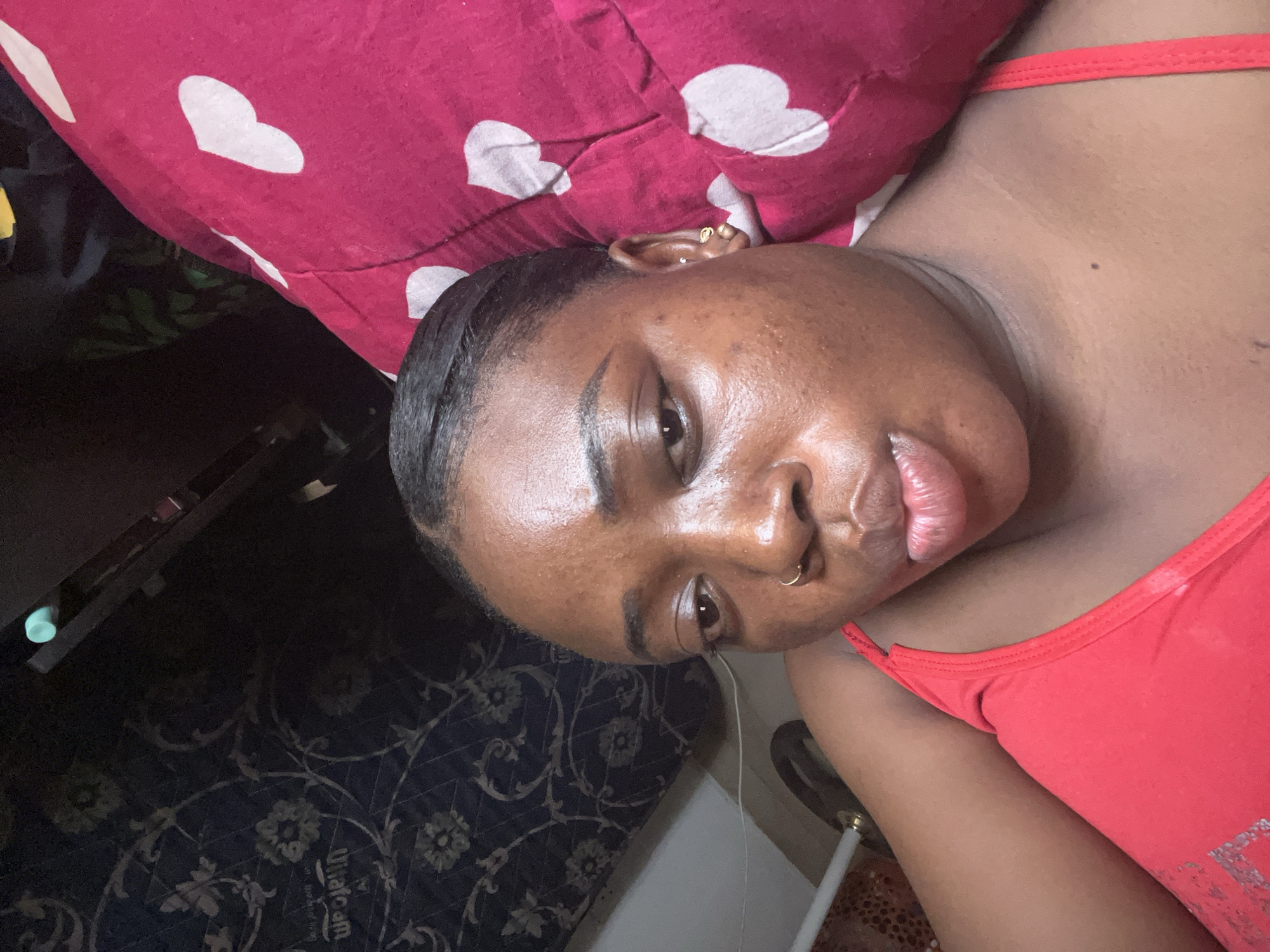 A person is lying down, with their head resting on a red pillow decorated with white hearts. The person has dark hair pulled back, dark eyes, and a nose ring. They are wearing a red tank top. The background shows a dark patterned rug and furniture. The image does not provide information to confirm the location is in Abuja, Nigeria.