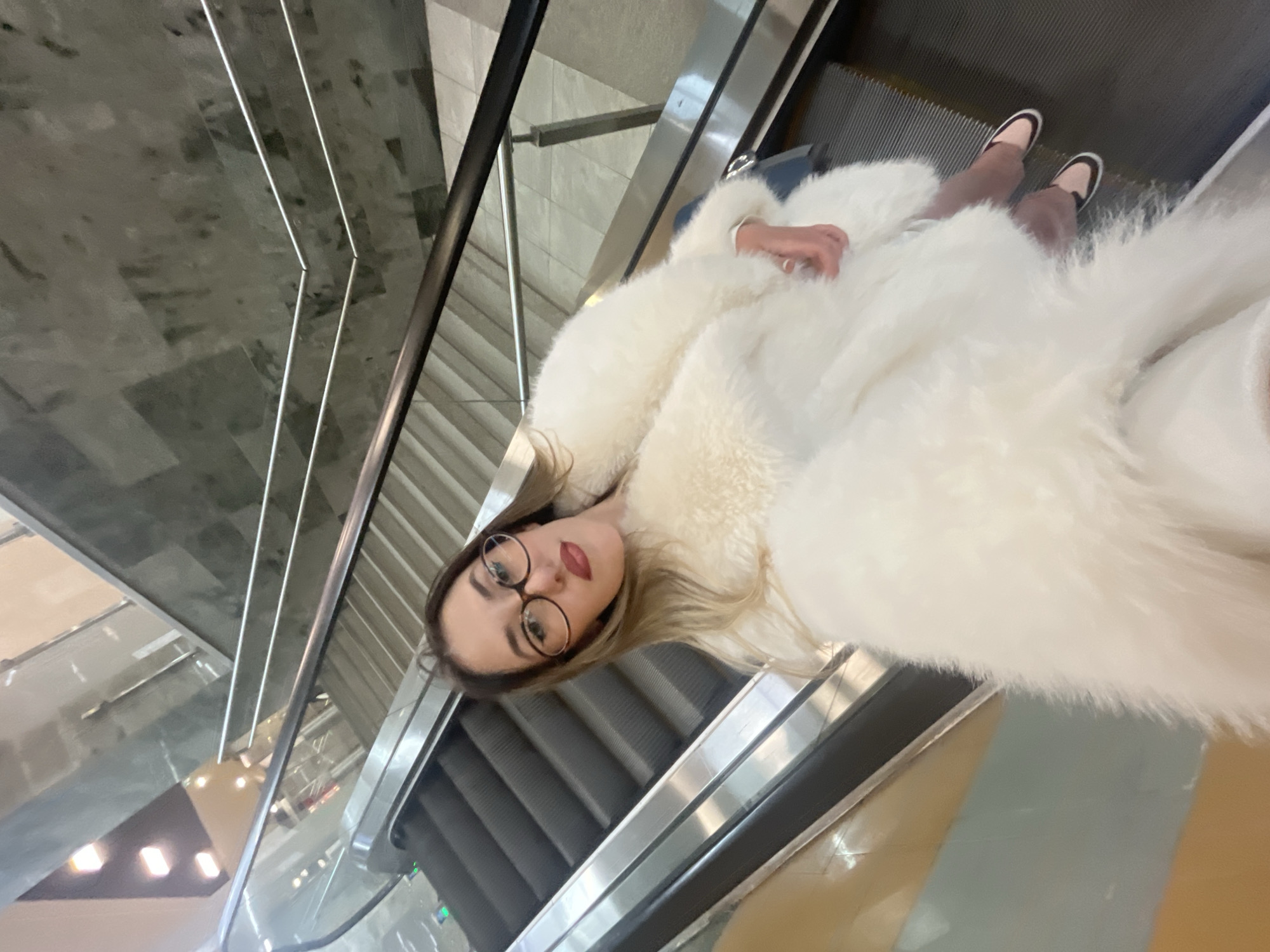 A young woman is depicted riding an escalator in a modern indoor setting, likely a shopping mall or public building in Sofia, Bulgaria. The image is taken from a slightly elevated perspective, looking down at her, or it is a selfie. She is wearing a voluminous white faux fur coat, round-framed glasses, and red lipstick. Her light brown hair is visible around her face. One hand is resting on her arm, revealing a ring on her finger. Her feet, clad in flat, light-colored shoes with black trim, are positioned on the escalator steps.
The escalator features black steps and metallic silver railings and side panels. The background reveals the architectural elements of the building, including geometric patterns on walls or ceilings, linear light fixtures, and reflective surfaces, possibly glass or polished metal, showing indistinct reflections of other parts of the interior and lights. The overall impression is one of a clean, contemporary environment. The woman looks directly at the camera with a neutral to slightly pleasant expression. There is no visible text in the image. The time of day is not discernible, given the indoor setting.