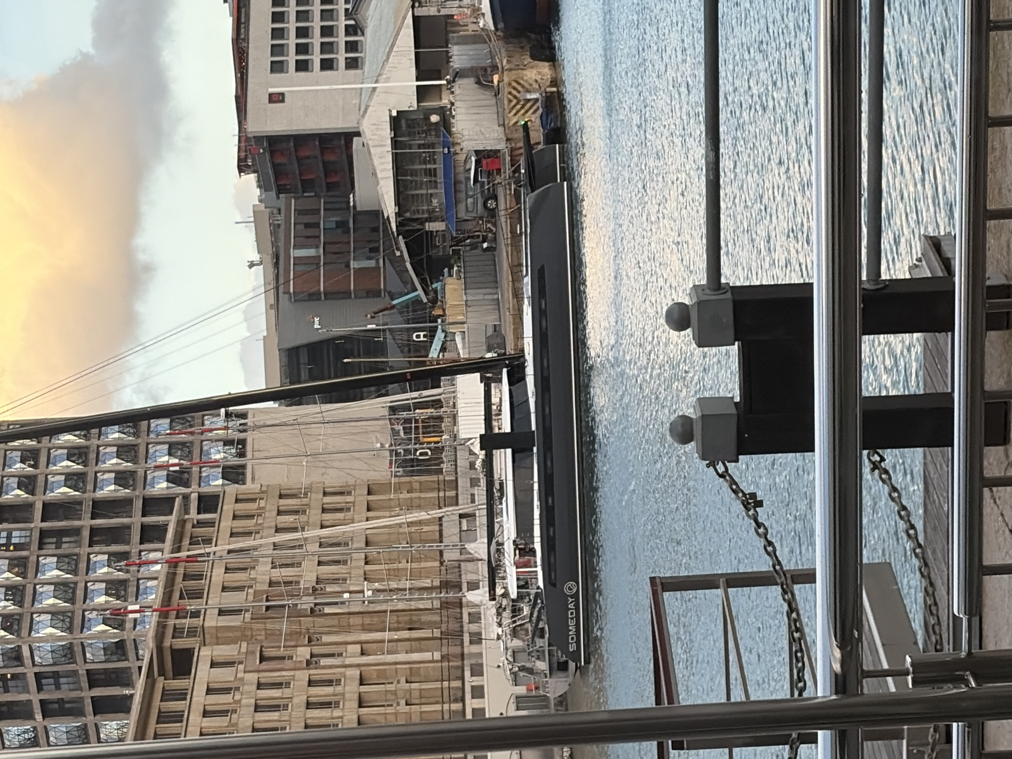 A long, sleek, dark vessel with the name "SOMEDAY" emblazoned on its side is docked in a harbor. The vessel runs nearly perpendicular to the viewer's perspective. Multiple buildings of varying heights dominate the backdrop. One building exhibits a unique, diamond-patterned facade. A crane towers above the construction site. A chrome railing and dark-colored structural supports frame the foreground. Water, likely from the harbor, reflects light. The location is Cape Town, South Africa, based on known architectural styles and harbor characteristics.