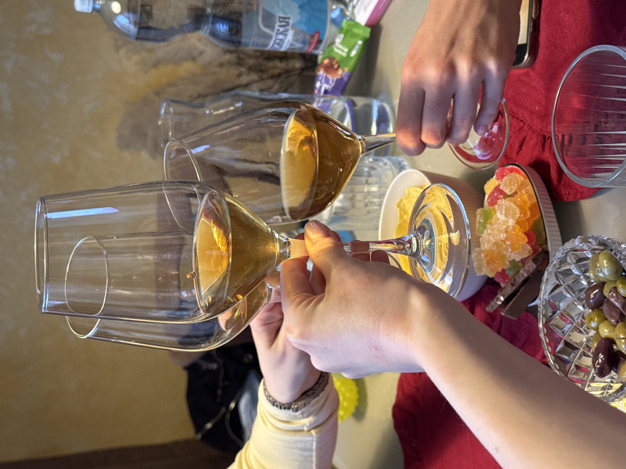Two people are toasting with wine glasses filled with a golden liquid. A plate of gummy candies and chocolates, a bowl of olives, and a small bowl of chips sit nearby on a table covered with a red cloth. In the background, there is a bottle of water and a snack package. The location appears to be indoors, but the specific city and location are not discernible.