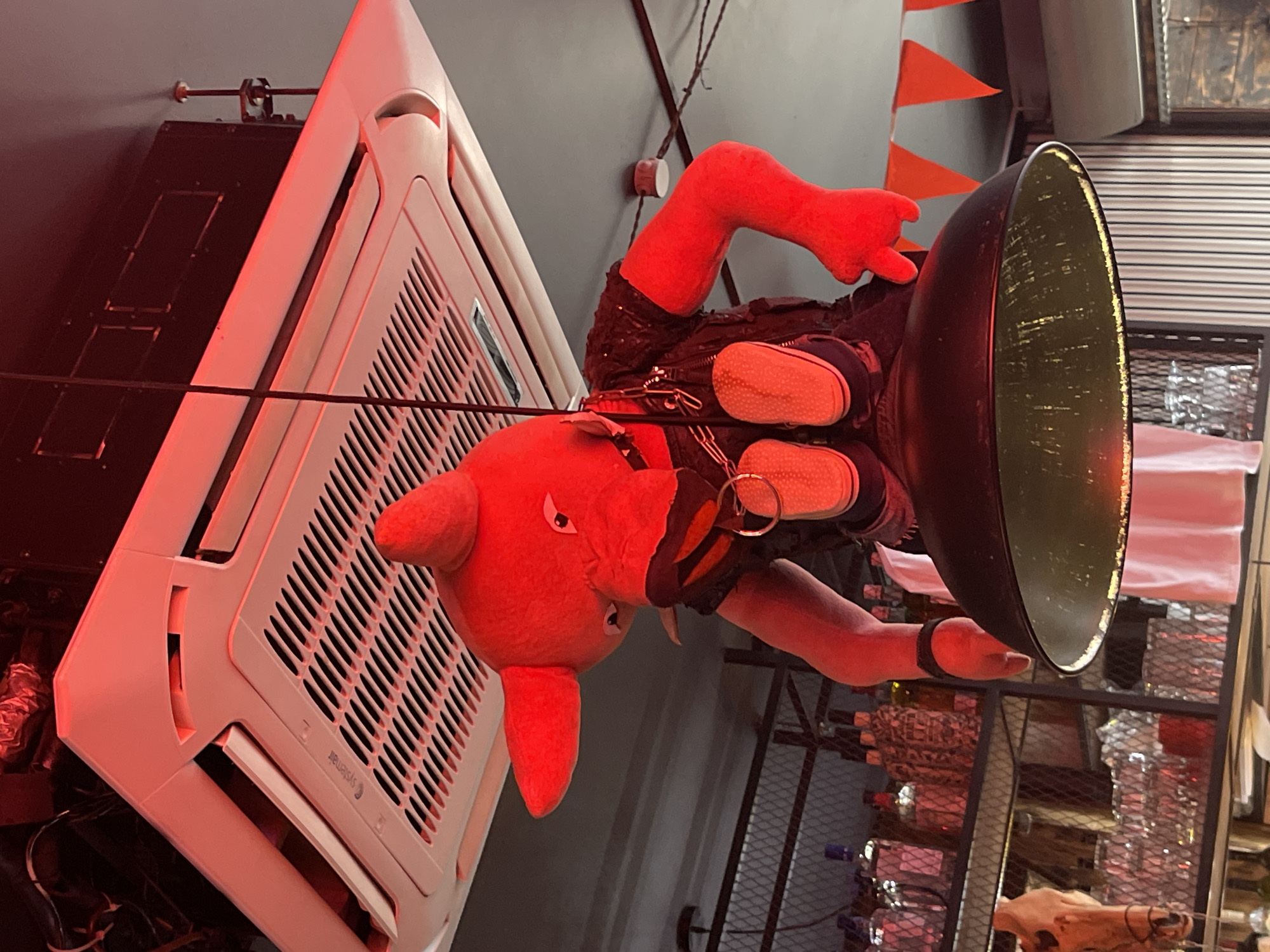 The image, taken in Saint Petersburg, Russia, depicts a surreal scene. An upside-down, bright red plush toy resembling a piglet in a studded leather outfit and small sneakers is suspended, possibly from a wire, against a gray wall. The piglet is positioned next to a large, white, rectangular air conditioning unit mounted high on the wall. A large, round, dark-colored lamp shade is attached to the piglet’s head. The background includes elements of a bar or restaurant setting, with visible shelves displaying various bottles, possibly alcoholic beverages. Additional details include orange triangular pennants hanging from the ceiling and a steer skull placed on a shelf. The lighting is primarily red.