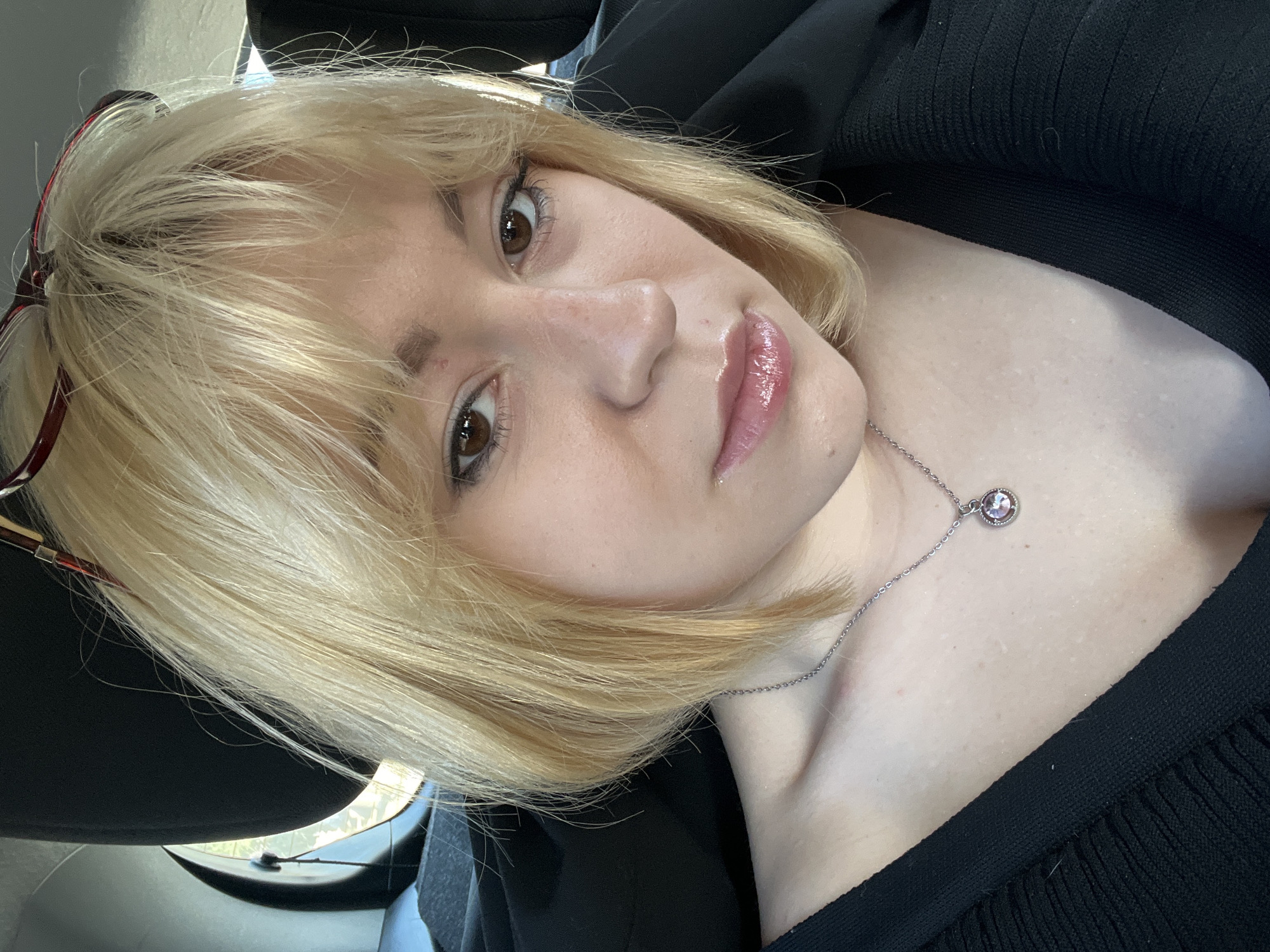 A fair-skinned young adult female with light blonde, short hair and brown eyes is the primary subject. She wears a black knit shirt and a silver necklace with a circular pendant. Part of a pair of eyeglasses with red rims is visible in her hair on the left side of the frame. She is looking directly at the camera, which is positioned at a slightly elevated angle relative to her. The background appears to be the interior of a car. The scene provides no identifiable link to Podlastva (Poljice), Montenegro.