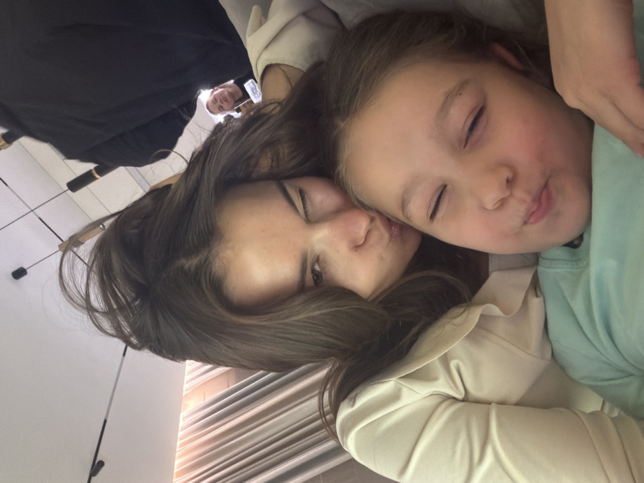 A woman and a young girl are closely snuggled together, both with their eyes closed and pursed lips as if kissing. The woman has long brown hair and is wearing a light-colored jacket. The girl has light skin and is wearing a green shirt. In the background, partially obscured, a man is looking directly at the camera while holding a phone.