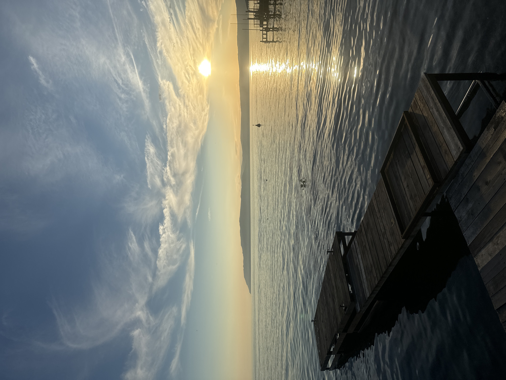 The image depicts a serene natural scene at sunset, potentially near Listvyanka, Russia, given the presence of a large body of water and distant hills that are characteristic of the region around Lake Baikal. In the foreground, a dark wooden pier extends into calm, rippling water. The pier appears aged, with visible planks and support structures.
The middle ground is dominated by the expanse of the water, reflecting the warm glow of the setting sun. A few small buoys or markers are visible on the water's surface, and a distant, silhouetted landmass or mountain range stretches across the horizon. The sun itself is a bright, glowing orb near the center-right of the frame, casting a shimmering reflection path across the water.
The sky above is a gradient of blues and yellows, transitioning from a clear, pale blue at the top to a warmer, golden hue near the horizon, infused with wispy, delicate clouds that are illuminated by the setting sun. These clouds create a soft, painterly effect across the sky.
In the background, to the right of the frame, another, more elaborate wooden structure, possibly a larger dock or a small building on stilts, is visible. The overall atmosphere is tranquil and picturesque, capturing the beauty of a lakeside sunset. No people are present in the scene.