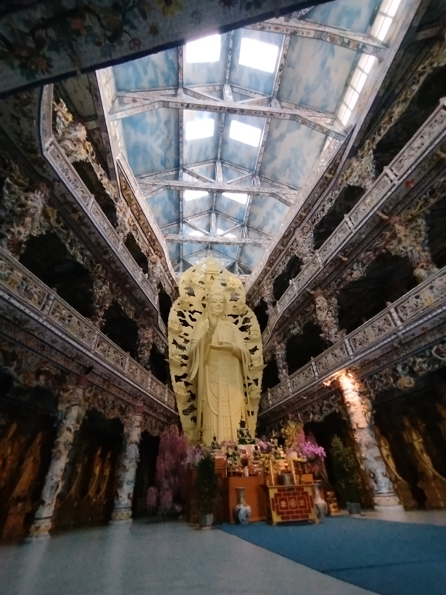 The image captures the interior of Bái Đính Temple in Xuân Thành, Vietnam. A large, light-colored statue stands prominently in the foreground, surrounded by flower arrangements and offerings on a table. The surrounding architecture features ornate, multi-tiered levels with dark, detailed carvings. A unique ceiling structure, possibly with skylights, is visible, with a blue and white sky-like design. The lighting is diffused and comes primarily from the skylights. The temple's floor appears to be blue in color. The viewpoint is from below, looking upwards towards the statue and ceiling.