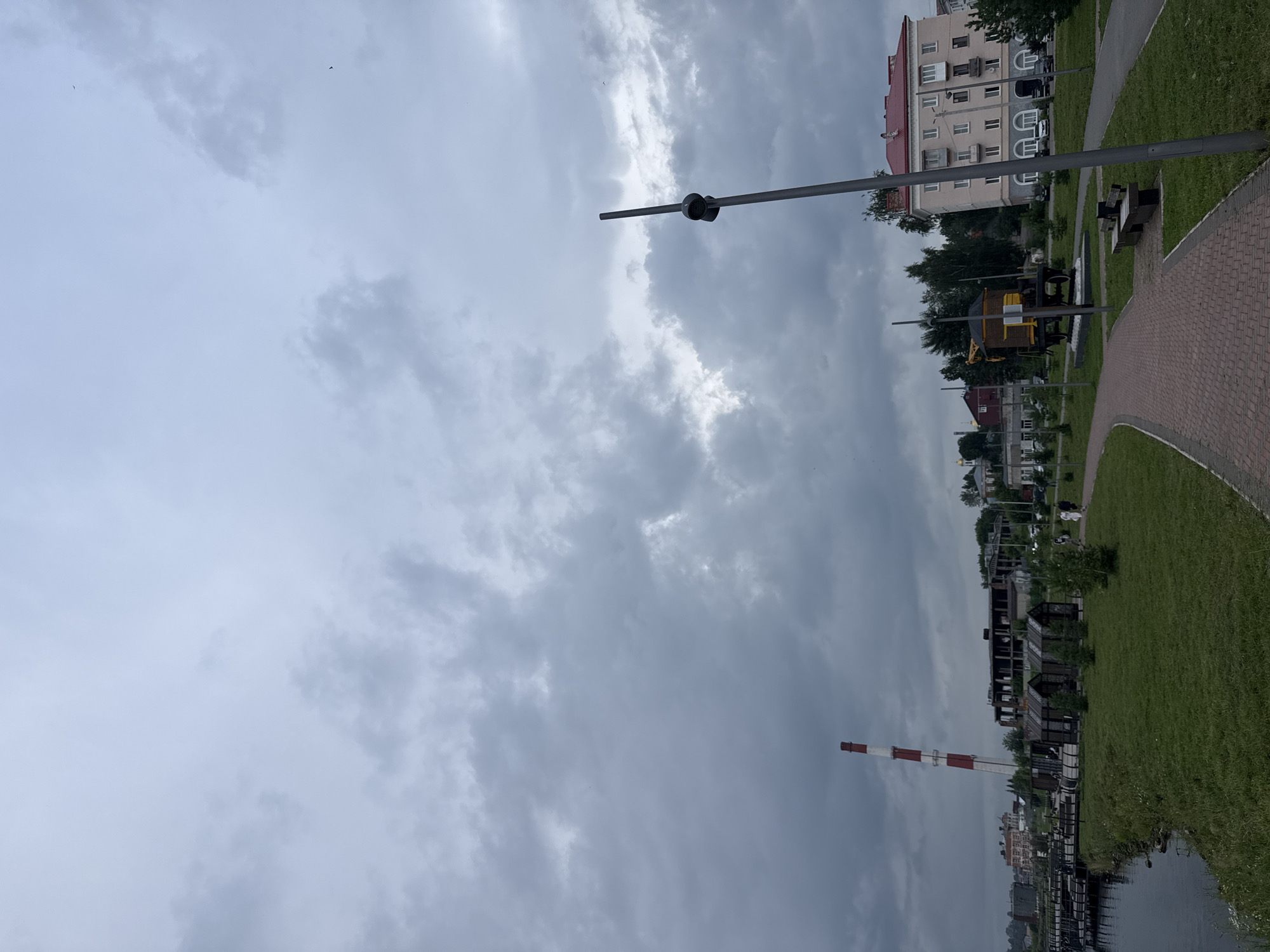 The image depicts an overcast day in the city of Lysva, Russia, likely near the waterfront. In the foreground, a brick pathway curves through a grassy area with several wooden benches. A tall, dark lamppost stands to the right of the path. Further back, a yellow and brown historical railroad car monument is visible. Beyond that, several small wooden structures are arranged on the grass. To the left, a body of water with wooden piers borders the grassy area. In the middle ground, a tall, red and white striped industrial chimney dominates the skyline, along with the skeletal remains of a large, unfinished building. Further in the distance, a multi-story apartment building with a red roof can be seen, along with other urban structures and trees. Two individuals are walking on the pathway in the middle ground. The sky is filled with heavy, grey clouds.