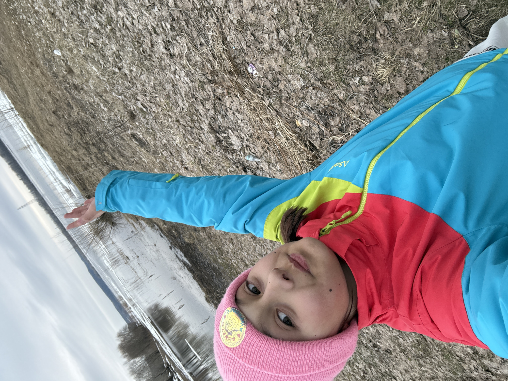 A person is lying on the ground near a body of water. The person is wearing a pink beanie, a blue and red jacket with yellow accents. The ground is covered with dry vegetation and some patches of snow. The body of water appears to be a river or lake with trees along the shoreline. The location is near a water body, and is likely in an outdoor environment near the city of Chaikovsky, Russia.