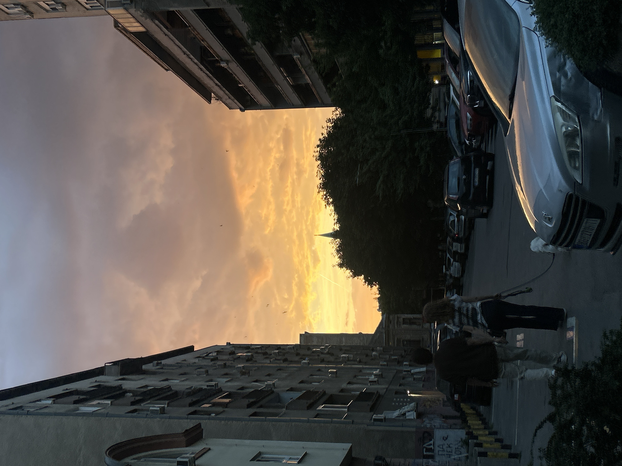 The image shows a street scene at sunset in Belgrade, Serbia. Buildings flank a street with parked cars on the right. Two people are walking on the sidewalk. The sunset is visible through a gap between buildings, with a church spire silhouetted against the orange sky. The time of day is early evening.