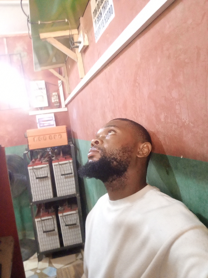 The scene depicts an interior room in Magboro, Nigeria. An adult Black male with short dark hair and a full beard, wearing a plain light-colored long-sleeved crew-neck shirt, is positioned in the right foreground. He is turned towards the right, gazing upwards and slightly to the right. The walls are painted reddish-brown, with a horizontal white trim visible along the right wall separating it from a darker, possibly green, lower section.
In the upper left background, a bright light source creates significant lens flare. Above this, a green panel is secured to the ceiling or upper wall with wooden brackets, showing exposed wiring. On the reddish-brown wall, a partially obscured, inverted vertical sign reads "DO NOT FIGHT HERE IN. ORDER BY POLICE."
To the left of the male subject, a power backup system is visible. This includes a metal rack holding four large, light-grey batteries with red tops, arranged in two tiers of two. Above these batteries, an orange inverter unit, partially labeled "GEECO", is mounted. Wires connect the system components. A small black fan is situated to the left of the battery rack. An electrical box is visible on the wall behind the inverter. The floor in the lower left consists of worn, patterned tiles.