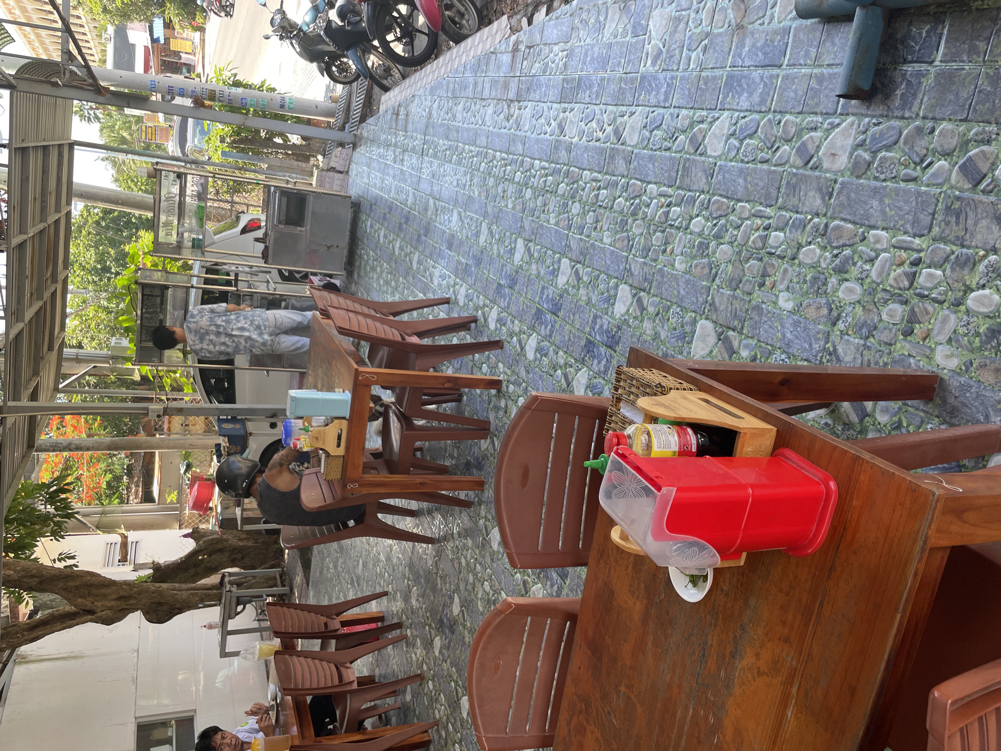 In Phu Thuy Ward, Vietnam, an outdoor dining area is depicted with multiple tables and chairs set up. A man wearing a black helmet and a dark sleeveless shirt sits at a table, facing away from the camera. Another person, partially visible, sits at a table in the background, wearing a yellow shirt and white pants. Several motorcycles are parked along a street to the right of the dining area. The ground is paved with cobblestone-like tiles, and a few trees provide shade. The scene appears to be a local eatery or a street-side restaurant.