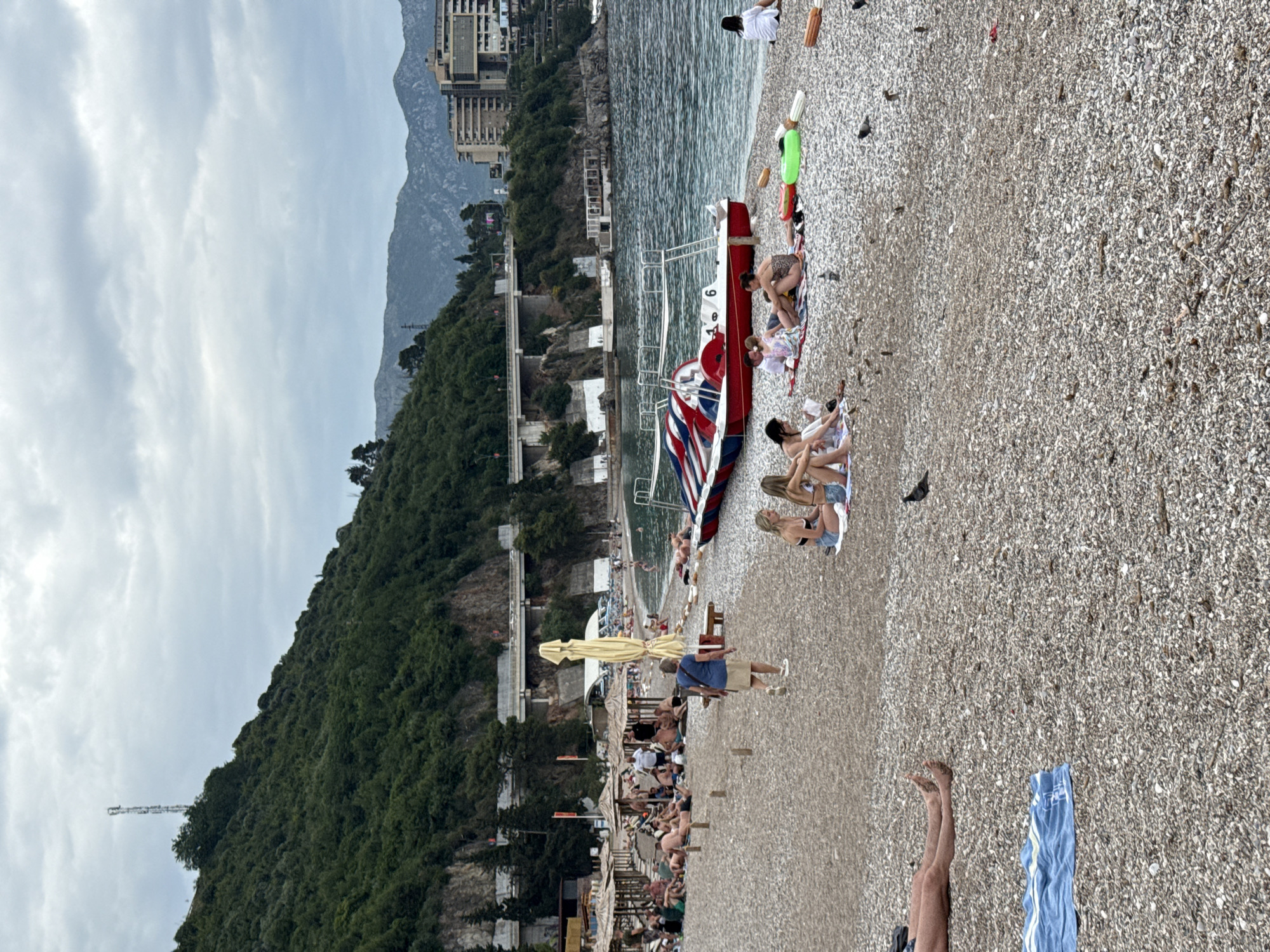 Here is a description of the image:
The image shows a pebble beach in Budva, Montenegro, filled with people sunbathing and relaxing. The beach is along a coastline with a backdrop of mountains and buildings. A red and white paddle boat with ladder rests on the beach. People are sitting on towels, and others are wading in the water. There are beach umbrellas visible along the beach.