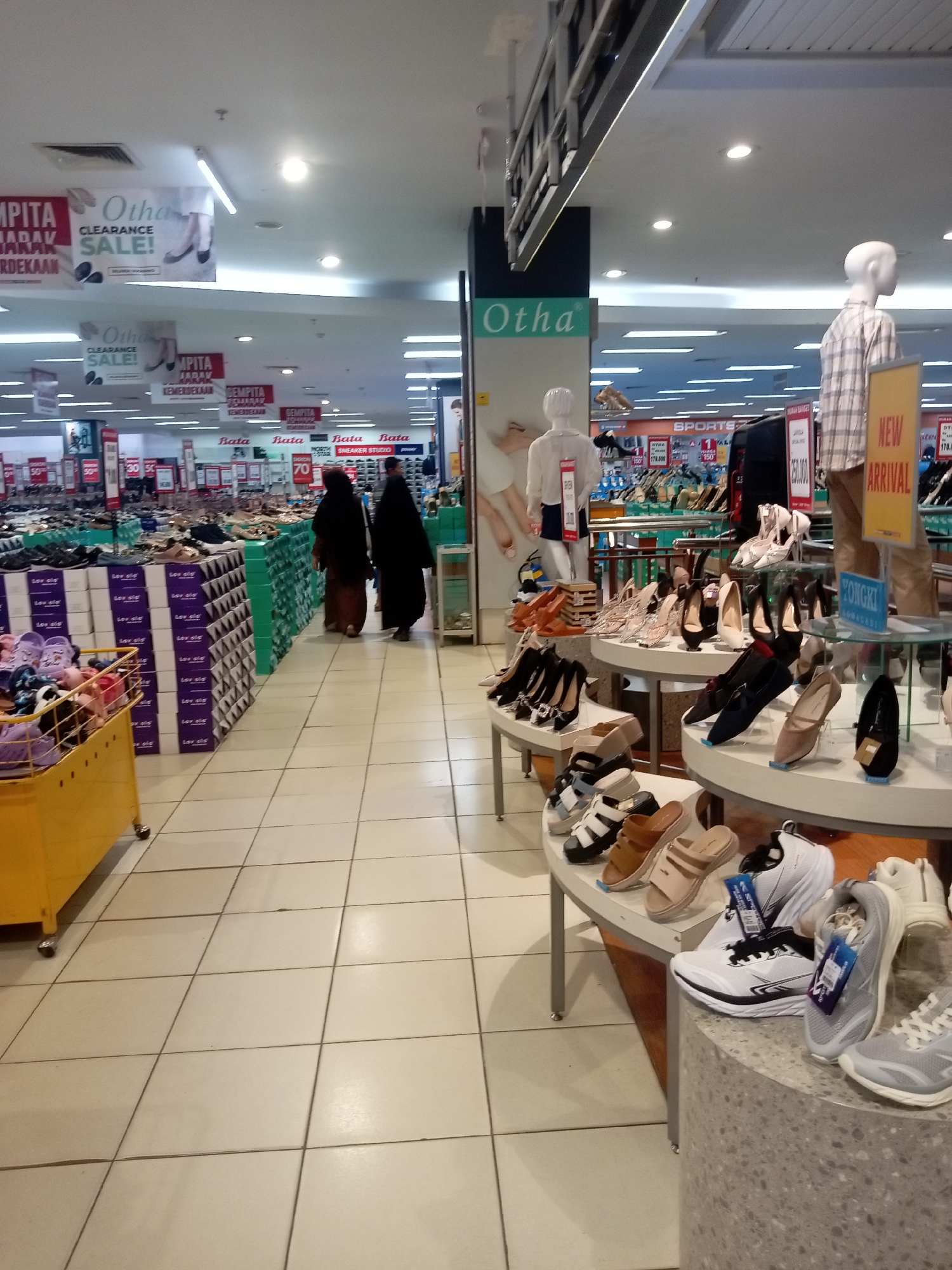 This image depicts the interior of a shoe store, likely a large department store or a specialized shoe retailer, located in Batam, Indonesia. The store appears to be well-lit with fluorescent lighting and features tiled flooring. Various displays of footwear are visible, organized on shelves and tiered stands. Among the displayed items are women's heels, sandals, and athletic shoes. Several mannequins are present, one dressed in a plaid shirt and another in a simple white top. Signs advertising "Otha" and "Bata" are prominent, suggesting these are likely brands sold in the store. There are also clearance sale banners visible. Two women, dressed in traditional Islamic attire covering their heads and bodies, are walking through the store, appearing to browse. In the foreground, a yellow shopping cart is filled with various shoes and footwear. The overall environment suggests a commercial setting focused on the sale of shoes.