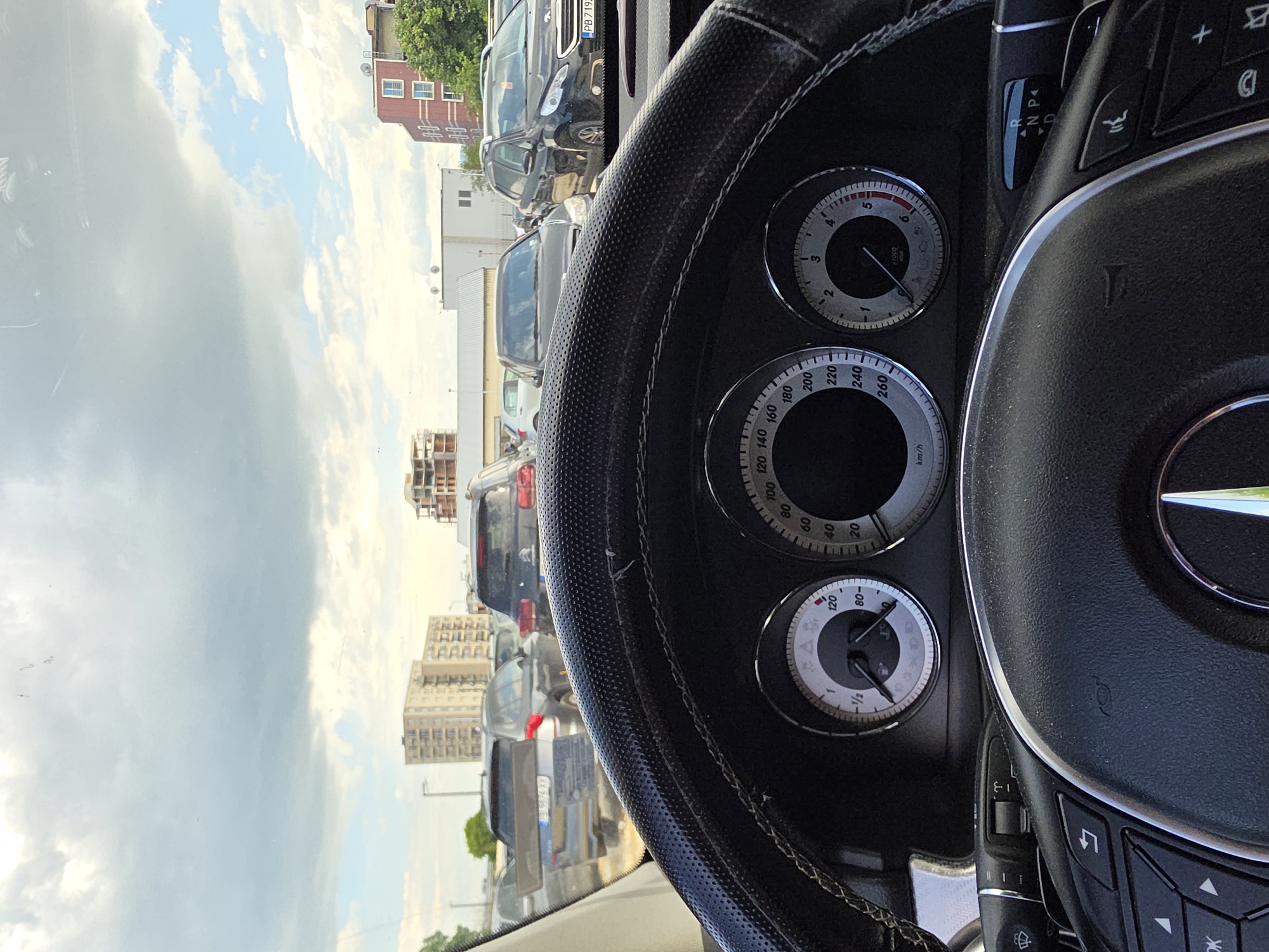 Here's a description of the image:
The image is taken from inside a car, focusing on the steering wheel and dashboard. The speedometer shows 0 km/h, and the fuel gauge indicates almost a full tank. Other dashboard instruments include a clock, and tachometer.
Outside the car, there's a line of parked cars. The sky is visible with scattered clouds. Buildings can be seen in the background. License plate "PB 7192" is visible on one of the cars. Based on the license plate and architecture, the location could be a parking lot in the city of Plovdiv, Bulgaria.