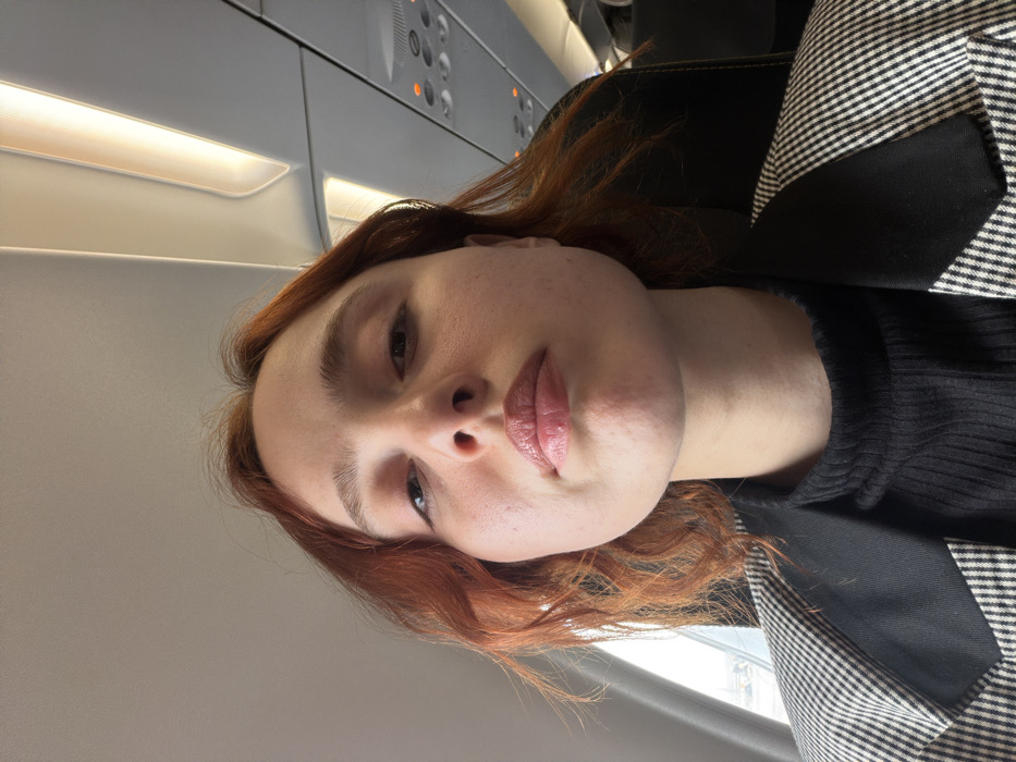 A young female with reddish-brown, shoulder-length hair is depicted in a close-up, upward-angled selfie taken inside a commercial airplane cabin. She has fair skin with visible pores, light brown eyebrows, and dark irises. Her lips are full and coated with a light pink or nude gloss, featuring a minor indentation on the upper lip. She wears a black ribbed turtleneck top underneath a black and white houndstooth or plaid blazer with solid black lapels.
Behind her, the dark headrest of a passenger seat is visible. To her left, a partial view through an aircraft window shows an overcast or hazy sky. Above her, the light grey airplane ceiling features a warm yellow-illuminated overhead lighting strip and a panel with multiple small, circular indicator lights, some of which are illuminated orange or red. The environment indicates the subject is aboard a commercial aircraft, in transit. Regarding the specified location of "Tuzla, Turkey," the aircraft is either at or in the vicinity of Sabiha Gökçen Airport (SAW), which serves the Istanbul area and is geographically close to Istanbul's Tuzla district, or within Turkish airspace.