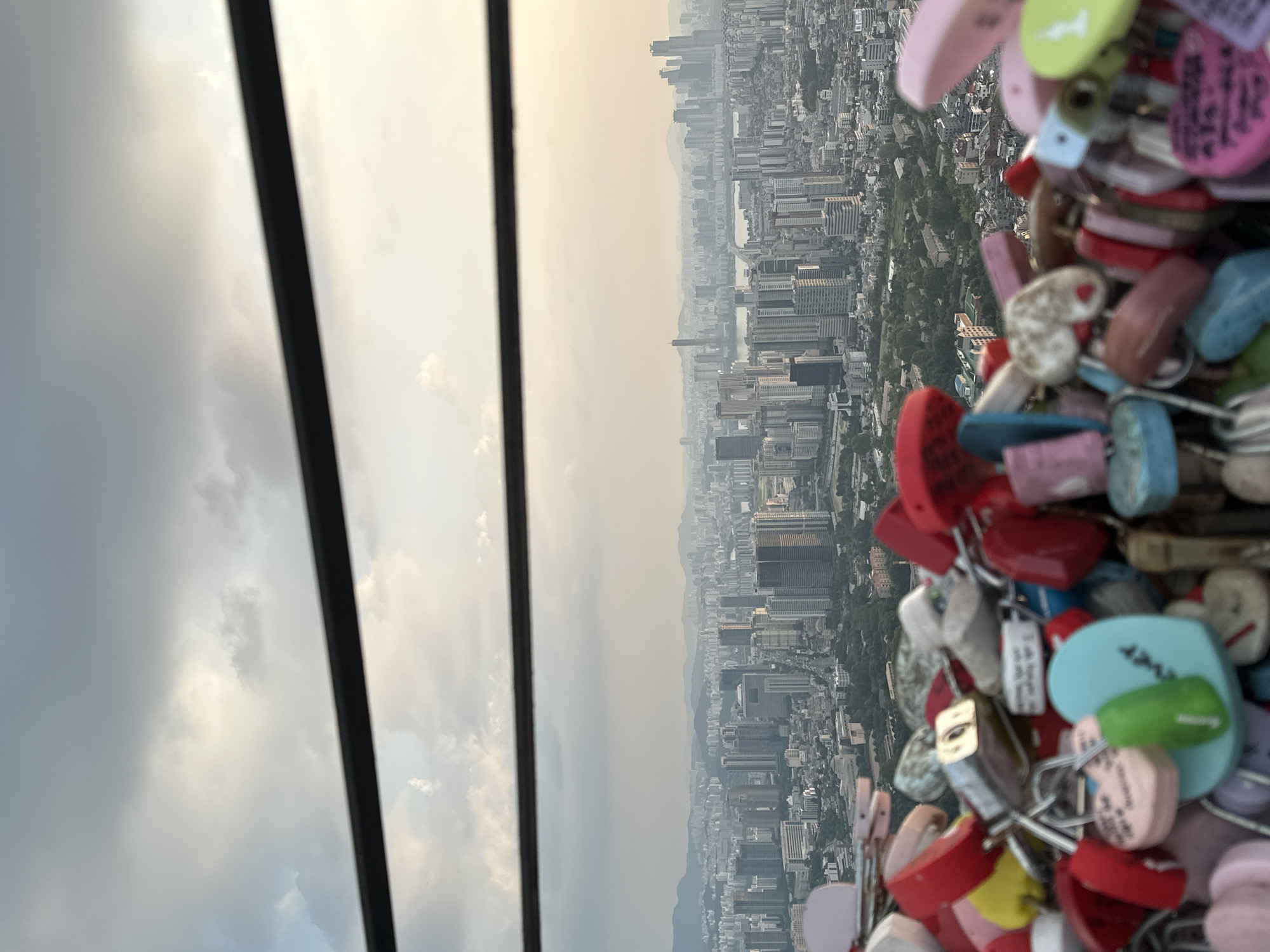 The image captures a close-up view of a collection of colorful love locks in the foreground, with a sprawling cityscape of Seoul, South Korea, visible in the background. The love locks, in various heart shapes and colors like red, pink, yellow, blue, and green, are densely clustered on a railing or fence. Some of the locks have inscriptions, and a few have keys attached.
The city view behind the locks reveals a vast urban landscape characterized by numerous high-rise buildings, residential blocks, and a hazy sky with scattered clouds. The distant horizon is marked by rolling hills or mountains. The presence of the love locks suggests a location like Namsan Seoul Tower, a popular tourist spot known for this tradition. The overall atmosphere is that of a scenic overlook in a major metropolitan area. There are no people visible in the frame. The image is taken from a slightly elevated perspective, looking down and across the city.