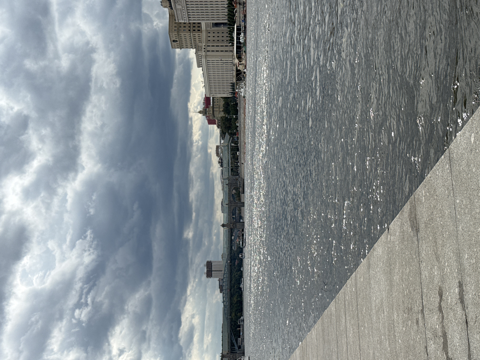 The image captures a wide-angle view of the Moskva River in Moscow, Russia, on a cloudy day. In the foreground, a concrete embankment lines the left side of the frame, with its textured surface showing some water marks. The river occupies the majority of the image, its surface shimmering with reflected sunlight, creating a dynamic texture. Several boats, including passenger vessels, are visible on the water.
In the background, a long bridge with a glass-enclosed walkway spans the river. Beyond the bridge, a collection of buildings forms the cityscape. To the right, a prominent, large, multi-story building with a light-colored facade and numerous windows dominates the skyline. Further to the left, another tall building with a distinctive silhouette stands out. The sky is filled with heavy, dark clouds, suggesting an overcast or potentially stormy weather condition, though patches of brighter sky are visible through breaks in the clouds. The overall scene depicts an urban waterfront with a mix of natural elements, architectural structures, and human activity.