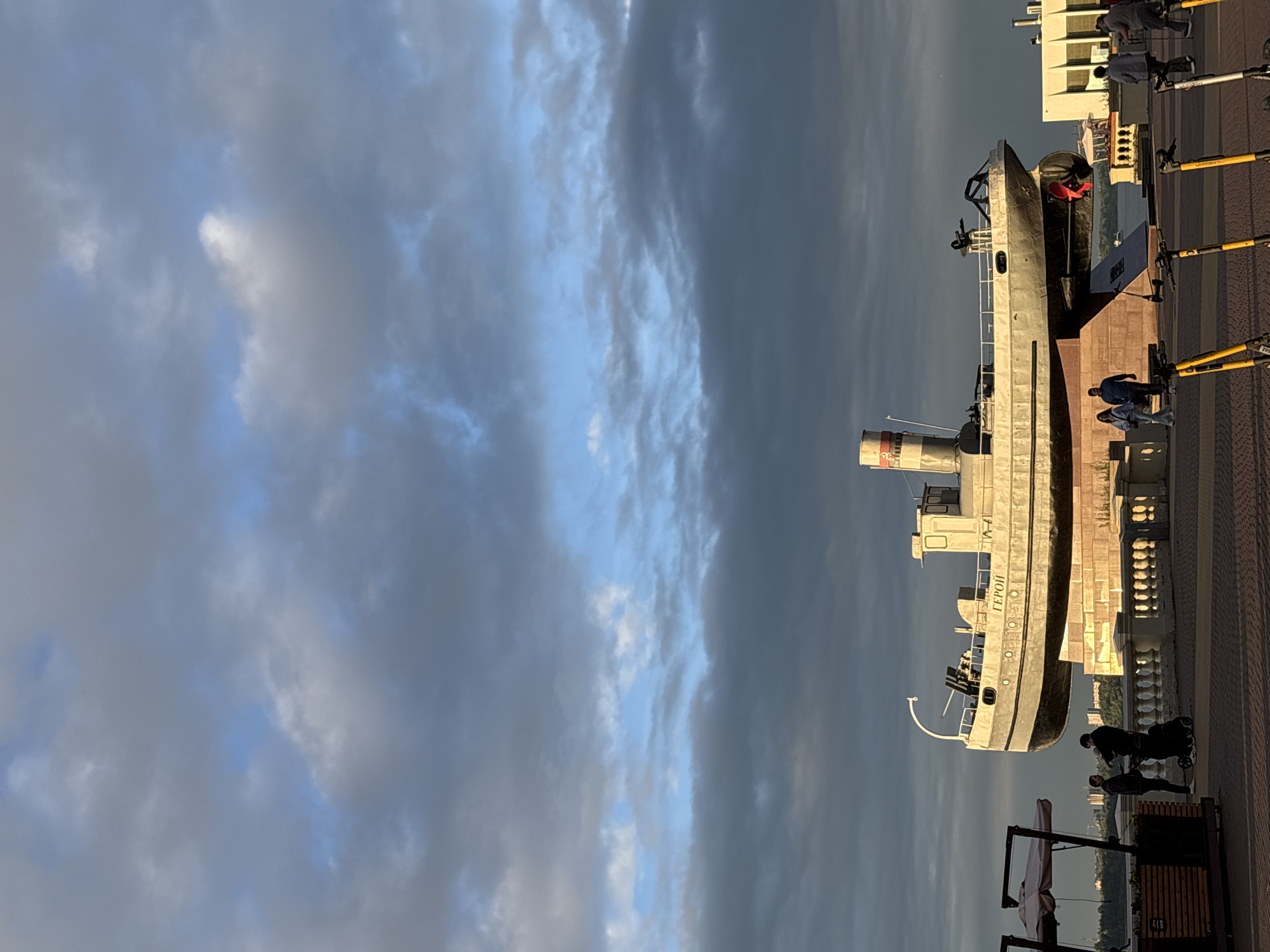 The image shows a monument of a ship, identified by the Cyrillic word "ГЕРОЙ" (Hero) on its hull, located in Nizhny Novgorod, Russia. The ship is mounted on a pedestal made of stone blocks, which is situated along a waterfront promenade. In the background, a cloudy sky with patches of blue can be seen. Several people are present on the promenade, including a couple pushing a stroller, another couple walking, and a group of people near a building in the distance. There are also several electric scooters parked in the foreground. The scene appears to be captured during the late afternoon or early evening, as indicated by the soft, warm lighting. The ship monument is a prominent feature, likely a historical or memorial site.