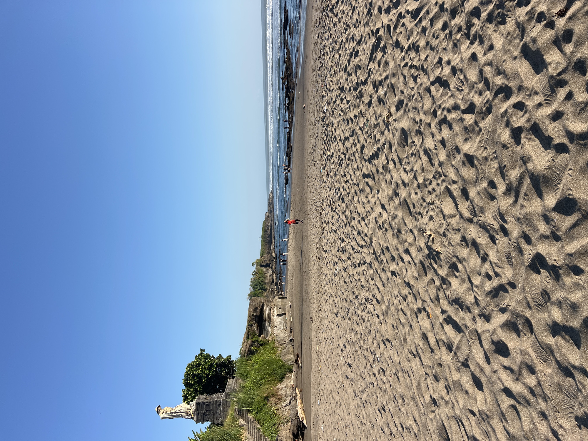 A wide shot captures a tranquil beach scene under a clear, bright blue sky. The foreground is dominated by coarse, light-brown sand, textured with the imprint of footprints and scattered debris. In the middle ground, a few individuals are visible. One person in a red shirt walks on the wet sand near the water's edge. Further out, a group of people are scattered near rock formations in the ocean, some appearing to be standing or interacting. The ocean itself is calm, with gentle waves rolling towards the shore. To the left, a clifftop overlooks the beach, adorned with a statue on a pedestal and lush green vegetation. A stone staircase leads up the cliff. The backdrop consists of more distant cliffs and the vast expanse of the ocean merging with the horizon under the unwavering blue sky. The image is a candid, eye-level view of a beach that does not appear to be in Kediri, Indonesia, as the architectural style and landscape are inconsistent with known locations in that city.