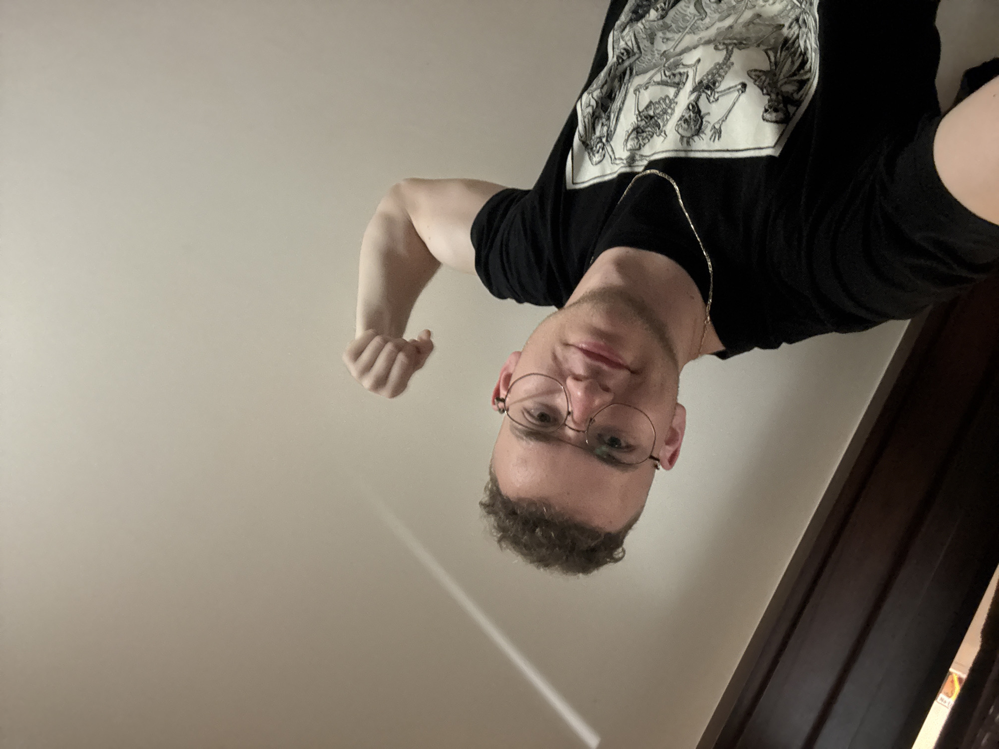 A man with curly hair and round glasses is angled downwards towards the camera. He is wearing a black t-shirt with a white graphic print and a gold chain. His right arm is raised and flexed. The background is a plain white wall with a diagonal shadow and a dark brown doorframe on the right. The location is not discernible from the image.