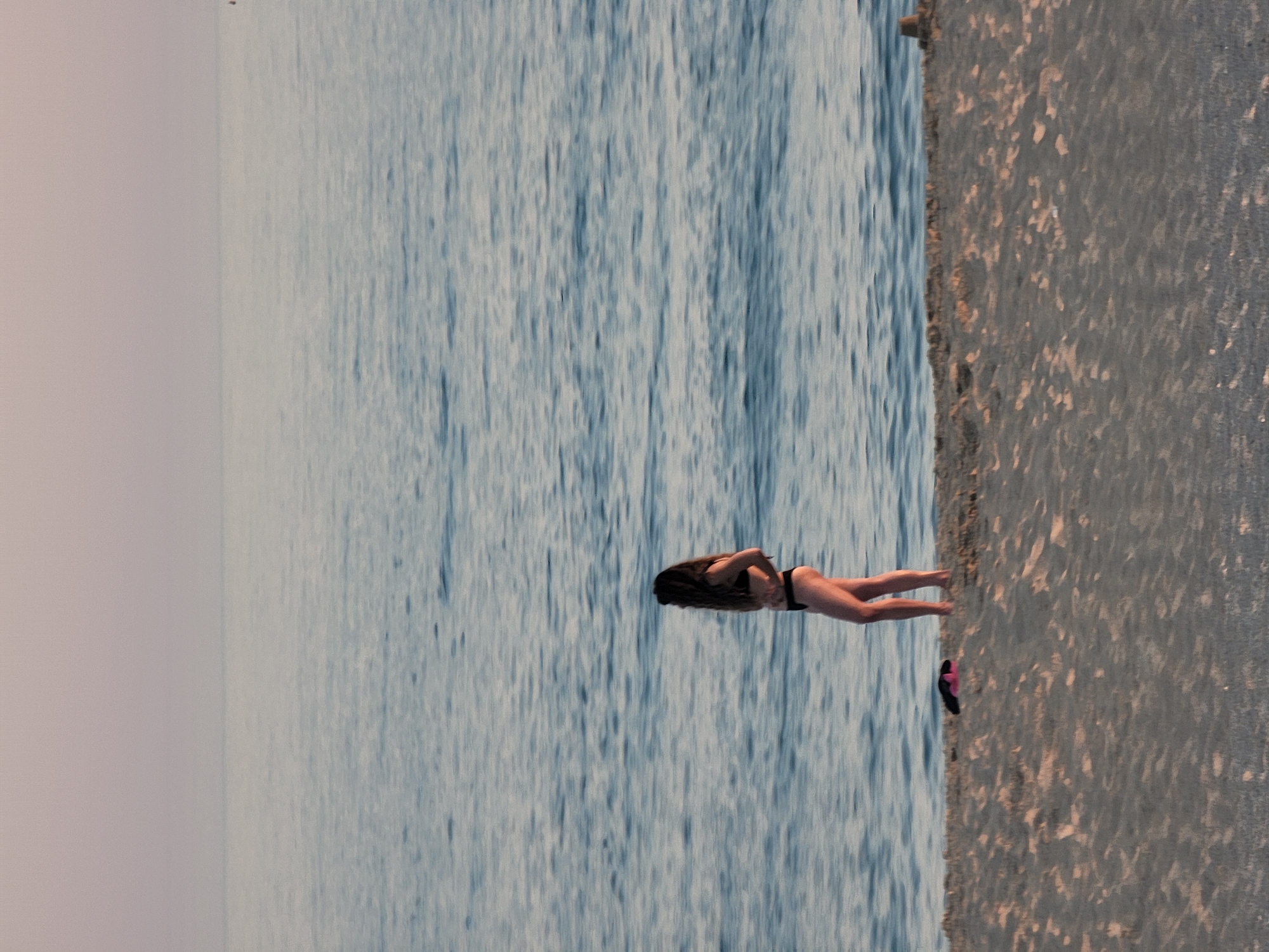 The image depicts a solitary woman standing on a sandy beach, facing the calm sea. She is wearing a black bikini and her long, dark hair is down. Her hands are placed on her hips. In the foreground, on the sand, are a pair of pink and black flip-flops. The sea is a muted blue with gentle ripples, and the sky above is a soft, hazy pinkish-orange, suggesting either sunrise or sunset. The overall impression is one of tranquility. This scene is consistent with the general coastal environment of Quarteira, Portugal, known for its beaches and seaside promenade.