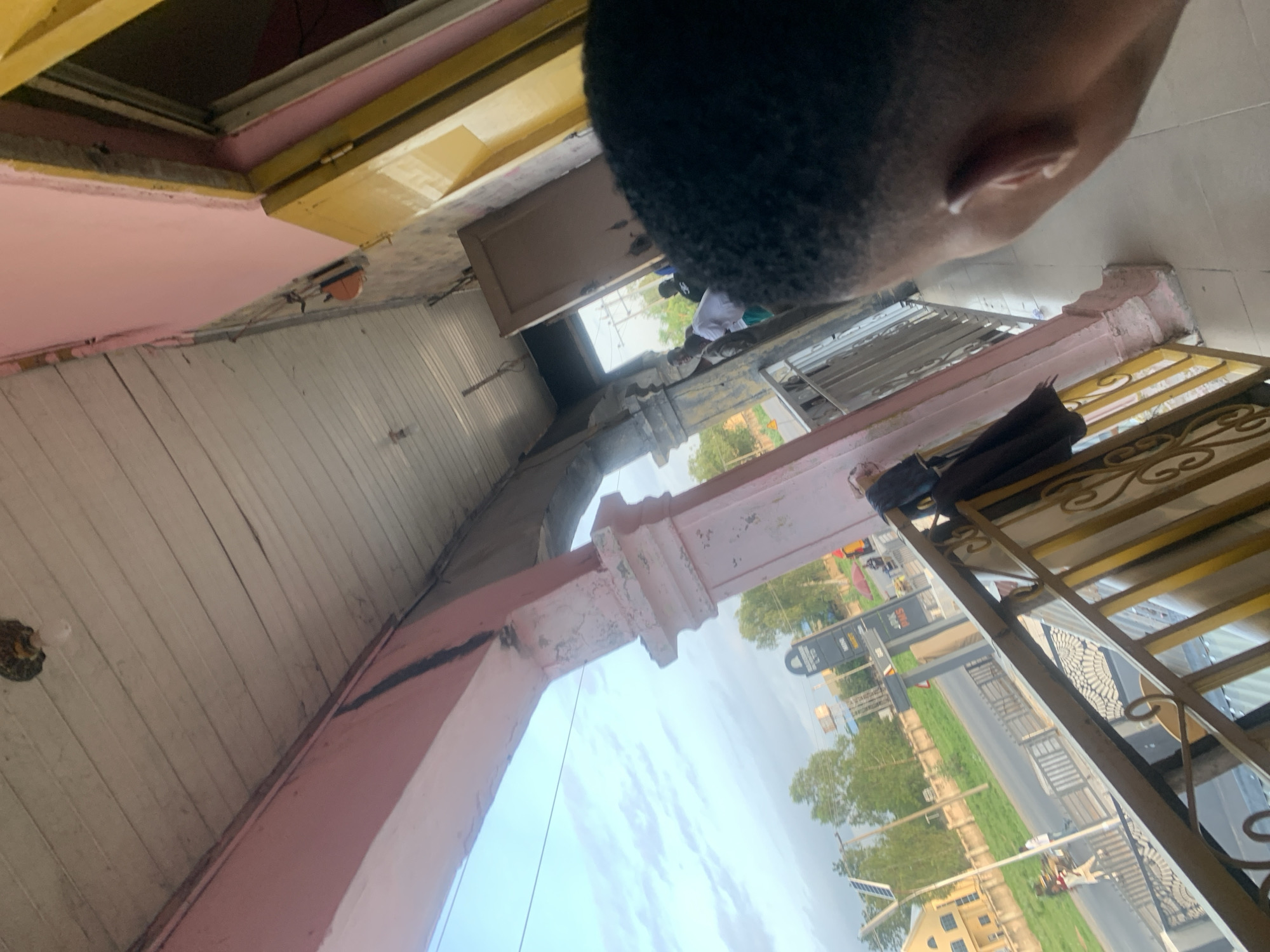 The image captures an elevated perspective of a street scene in Jalingo, Nigeria. In the foreground, a person's head and shoulder are visible, facing away from the camera. The person has dark, short, curly hair. The view extends to a covered walkway or balcony with a pink-painted railing and a wooden ceiling. A light fixture is attached to the ceiling. Beyond the walkway, a street is visible with a yellow building on the left and a large signpost with text, possibly advertising businesses. Several trees line the street, and the sky is partly cloudy. A few individuals are visible on the street and within the covered area, some walking and others standing. The overall impression is of a bright, daytime scene in an urban environment.