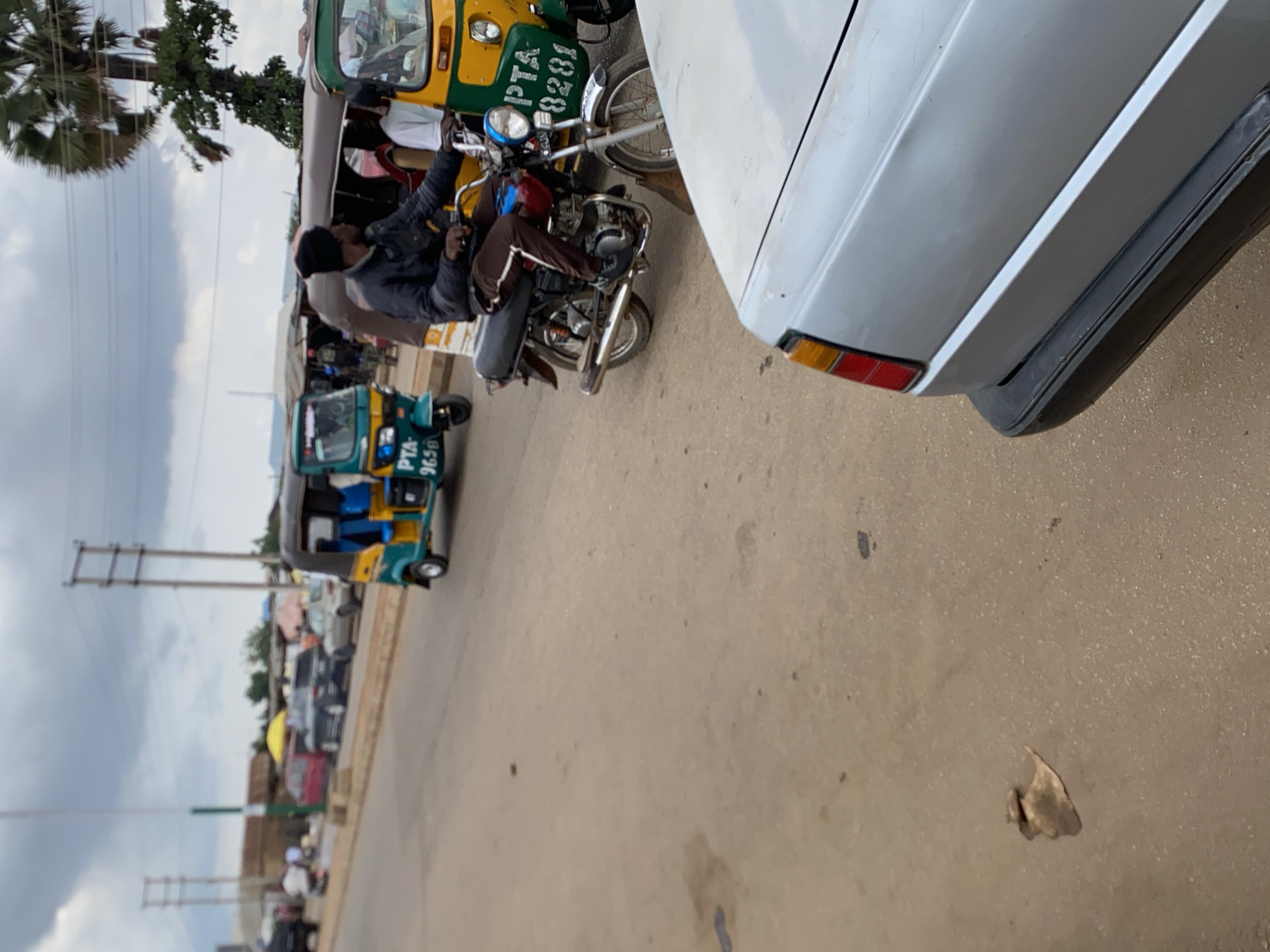 This is a medium-shot, eye-level, natural-light photograph taken on a street in Bukuru, Nigeria. The foreground is dominated by the rear quarter of a silver car, its black bumper and taillight assembly visible. The paved road surface shows some staining and debris, including a solitary leaf-like object.
In the middle ground, a man wearing a black beanie, black jacket, and brown pants rides a motorcycle, which is painted with red and blue accents on the fuel tank and a silver exhaust pipe. The motorcycle is positioned in front of a yellow and green tuk-tuk, identified by the license plate "PTA 8281". Another blue and green tuk-tuk, with license plate "PTA 9658", is visible to the left.
The background depicts a street scene with several other vehicles, including a red car and a dark-colored SUV, parked or moving along the road. A yellow umbrella is open, suggesting a market stall. A palm tree with sparse fronds is visible on the right side of the frame. Overhead, utility poles and power lines crisscross the cloudy sky. The overall impression is of a busy, somewhat dusty, urban environment.