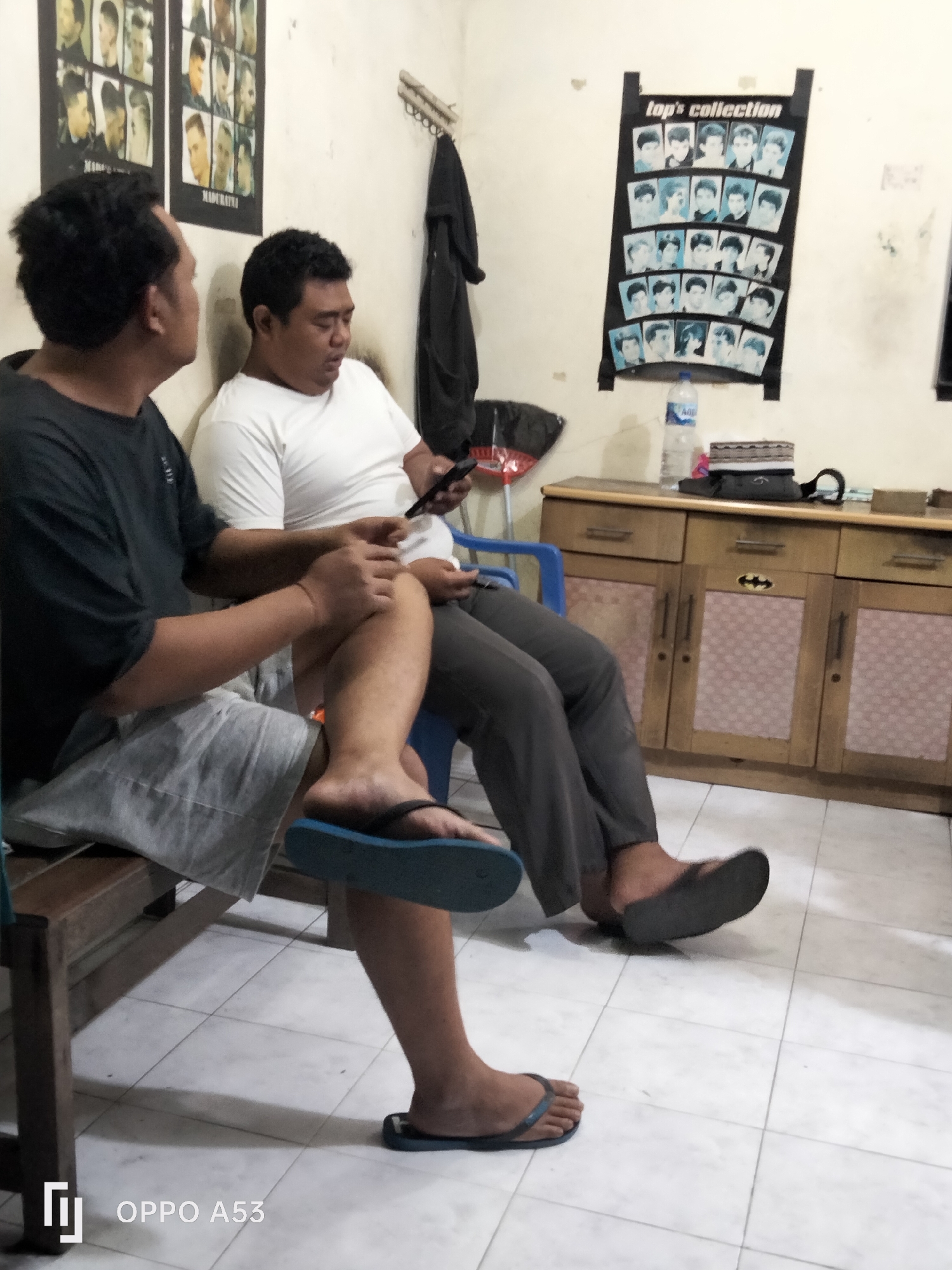 The image depicts two men sitting in what appears to be a barbershop or a similar establishment in Denpasar, Indonesia. The man on the left is wearing a black t-shirt and grey shorts, with a blue flip-flop on his left foot. He is looking towards the man on the right. The man on the right is wearing a white t-shirt and grey pants, and is holding a mobile phone in his right hand. He is looking down at the phone.
Behind the men, on the wall, are two framed posters showcasing hairstyles. To the right of these posters, a black garment hangs on a hook. Further to the right, there is a wooden cabinet with drawers and doors. On top of the cabinet, a bottle of water, a religious cap, and other items are placed. The floor is tiled with a white grid pattern. The overall environment suggests a casual, indoor setting.