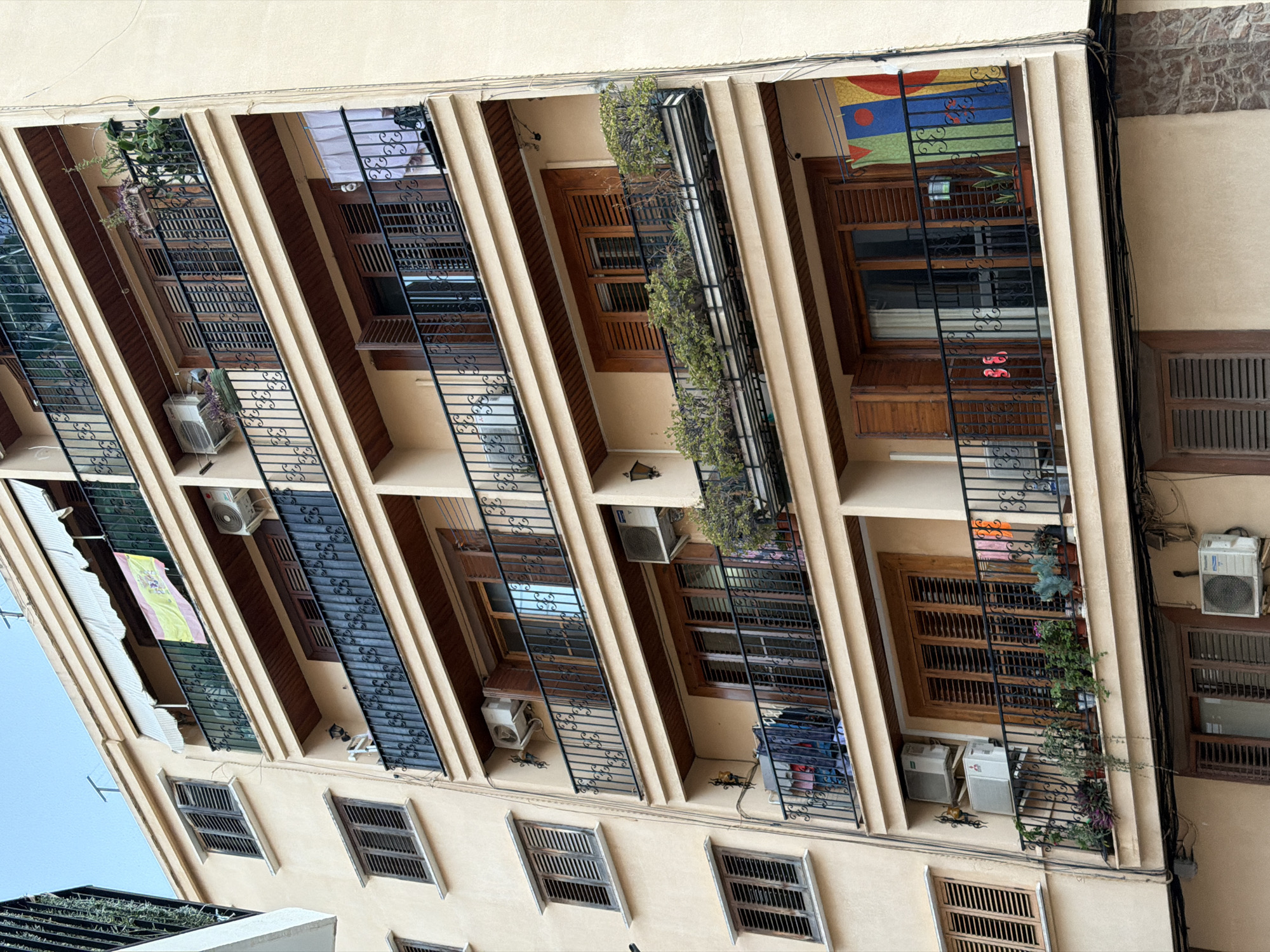The image shows a facade of a multi-story apartment building. The building has a light beige exterior and features multiple balconies with black wrought-iron railings. Each balcony has wooden shutters on the windows. There are several air conditioning units mounted on the exterior walls. Various items are visible on the balconies, including plants in pots and hanging baskets, laundry hanging on a line, and some folded items. A Spanish flag is prominently displayed on an awning above one of the balconies. The building is situated in an urban environment, as suggested by the view of the sky and the architecture. The specific location is Benimàmet-beniferri, Spain.