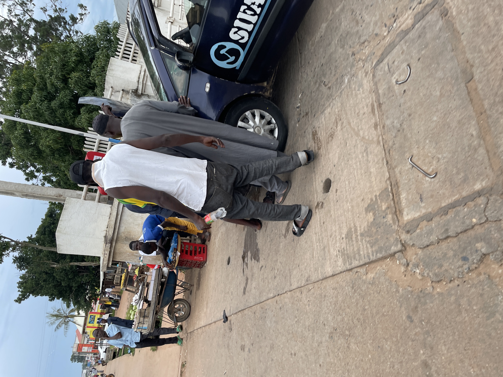 The image depicts a street scene in Jos, Nigeria, with a dark blue vehicle parked on the right side of the frame. The vehicle has a logo on its side that appears to read "SIFA". Several people are interacting near the vehicle and on the sidewalk.
In the foreground, a man wearing a white sleeveless shirt and dark pants is holding a clear plastic bottle. Another person, partially visible in gray attire, stands near the car door. A third individual in a blue shirt and yellow shorts is bent over a red crate filled with items, possibly goods for sale. A blue wheelbarrow filled with green produce, likely melons, is positioned behind the red crate.
Further back, a man in a light blue shirt and dark pants stands on the left side of the street, looking towards the left. Other individuals are visible in the background, near market stalls or shops. The environment appears to be an outdoor market or roadside commercial area, with trees and buildings visible in the distance. The ground is paved with concrete and appears somewhat worn and stained. The sky is overcast.