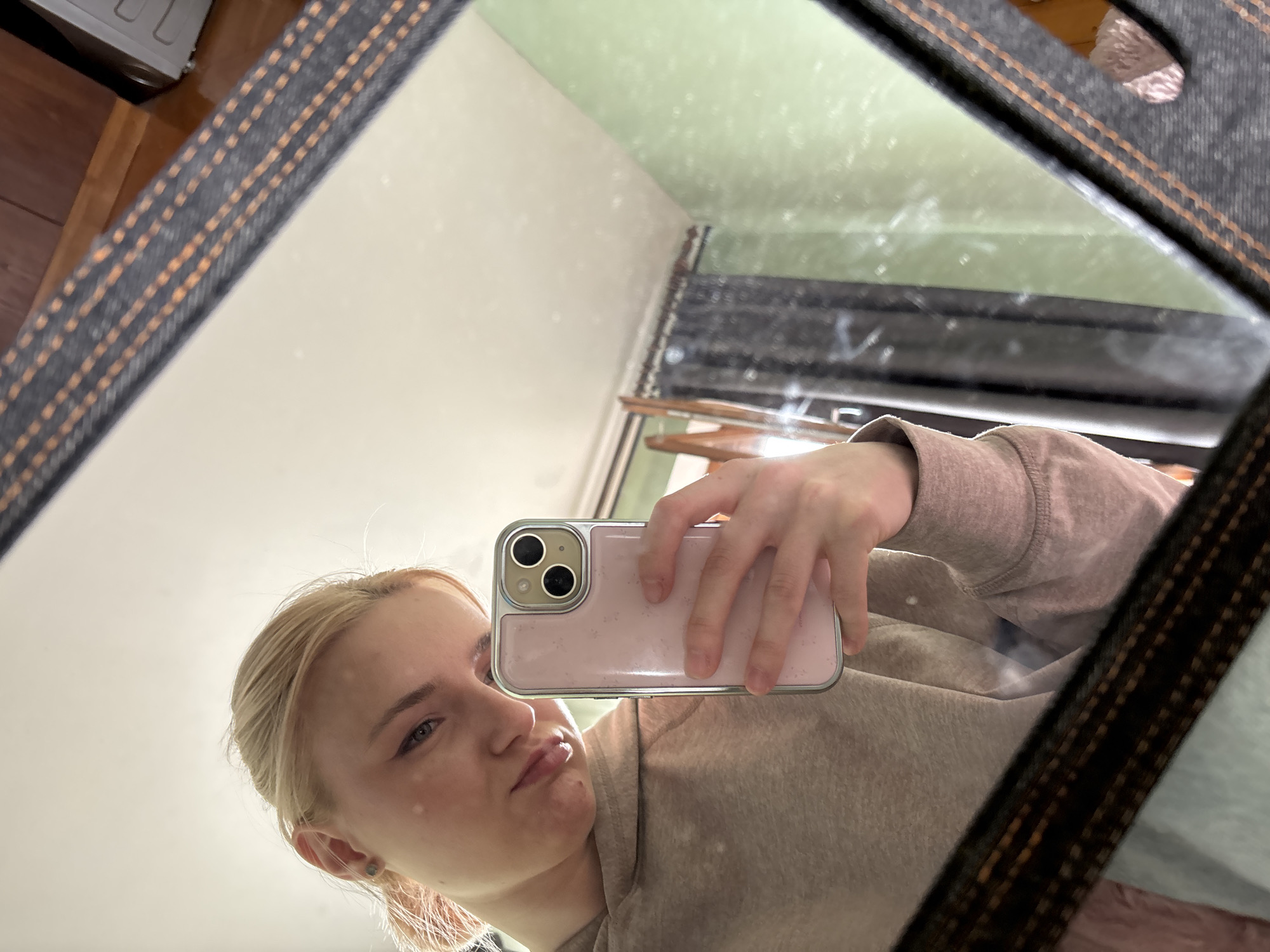 A young blonde woman with fair skin and light makeup takes a selfie in a mirror. She is wearing a light brown hooded sweatshirt and has a pink phone with a dual camera in her right hand. Her hair is pulled back in a ponytail. The mirror is framed with dark material featuring orange stitching. In the reflection, a green wall, a window with dark curtains, and part of a wooden dresser are visible. The scene appears to be indoors, likely in a residential setting. The location is not specified beyond "Unknown, Russia."