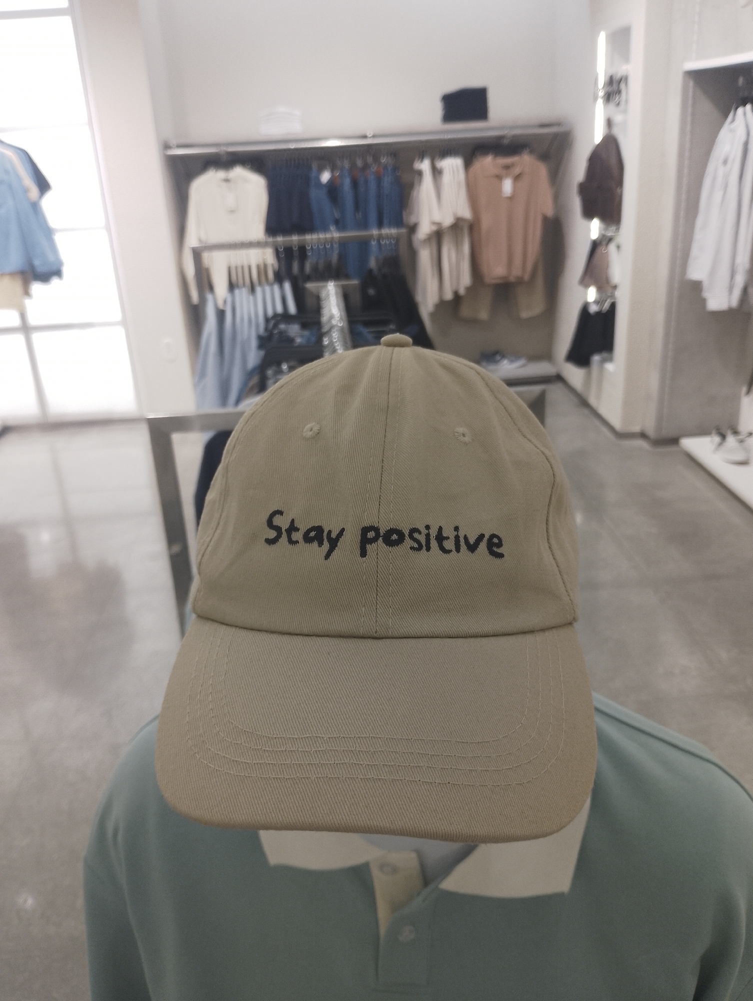 A tan baseball cap with the words "Stay positive" embroidered in black lettering is placed on a mannequin's head. The mannequin is wearing a light blue collared polo shirt. The background shows a retail clothing store with various clothes hanging on racks. The location cannot be determined from the image.