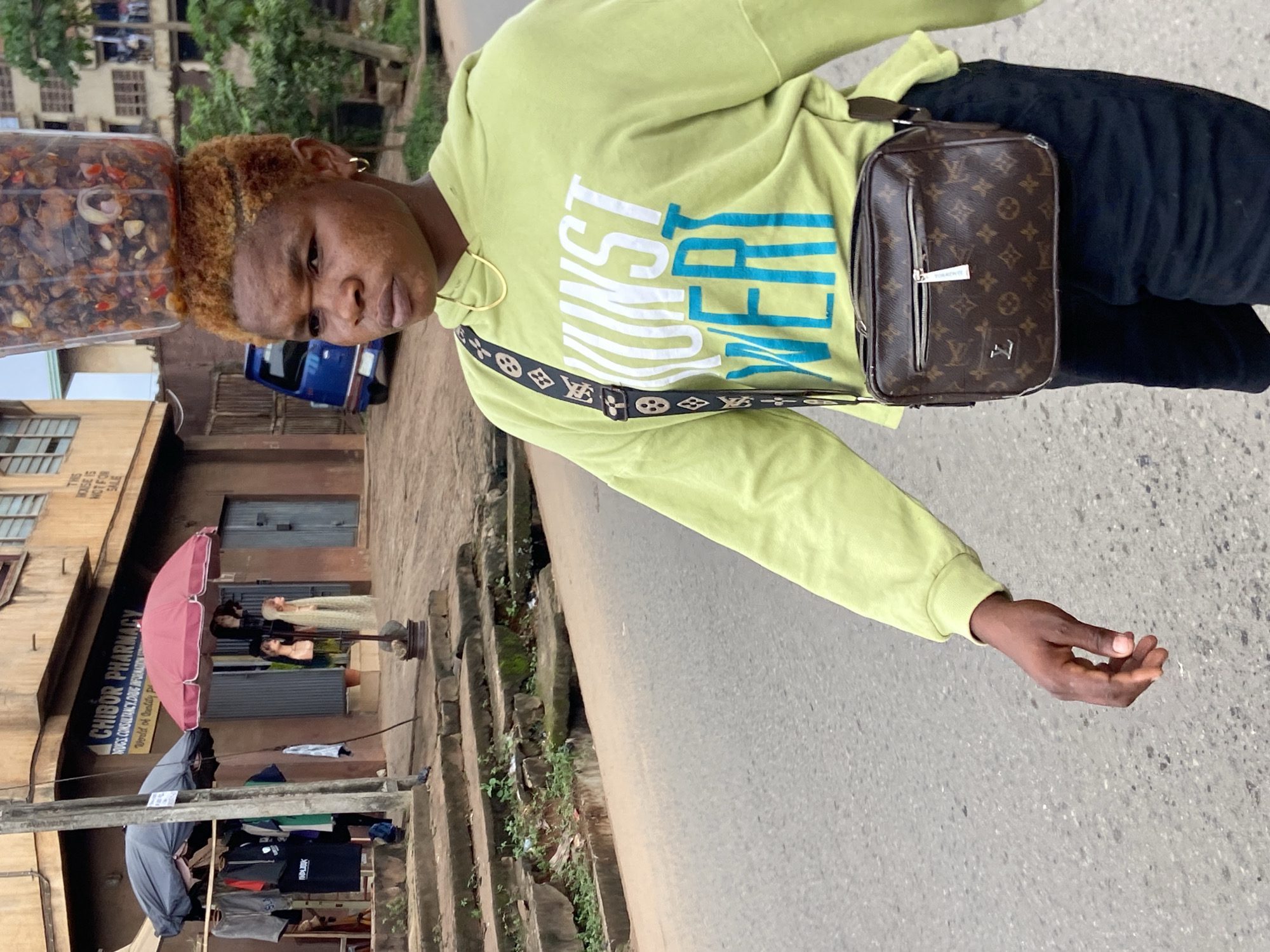 In Nkpor, Nigeria, a young woman with short, dyed blonde hair walks down a paved street. She is carrying a clear plastic container filled with food items on top of her head and wearing a lime green hooded sweatshirt with white and blue lettering that reads "KUNST WER". A brown, monogrammed crossbody bag hangs diagonally across her chest. She is also wearing black shorts and a gold chain. In the background, a blue vehicle is parked, and shops line the street. One shop, "CHIBOR PHARMACY," is visible with displays of clothing and mannequins outside. The street appears to be in a commercial area with buildings and sidewalks.