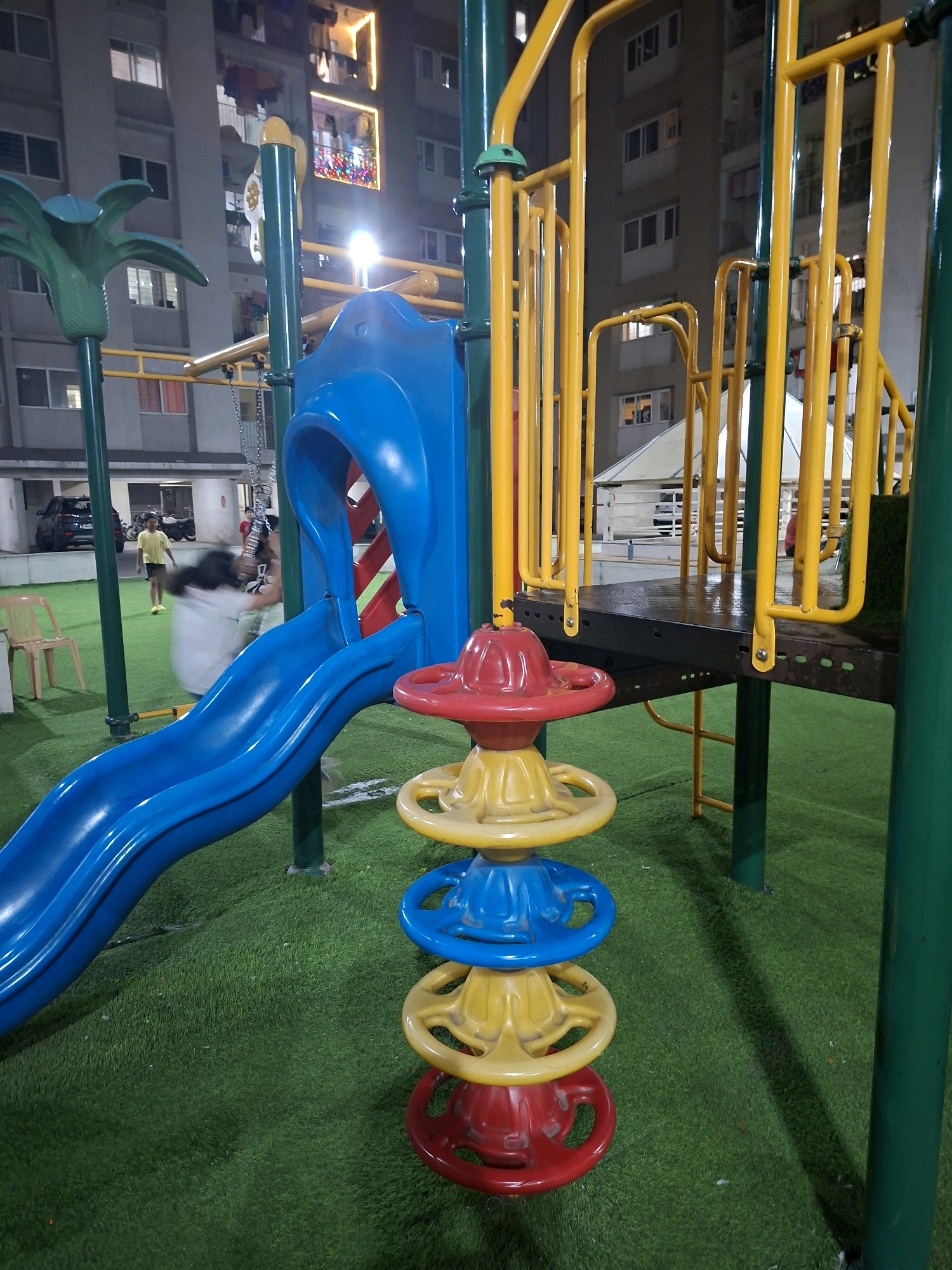 The image depicts a children's playground at night, located in Pune, India. The playground features a blue slide, yellow railings, and green artificial turf. In the background, a residential building with illuminated windows is visible. Two people, one wearing a yellow t-shirt and the other a dark top, are present in the playground. The person in the dark top appears to be in motion, possibly swinging or running, causing a blur. A red, blue, and yellow spinning toy is positioned in the foreground.