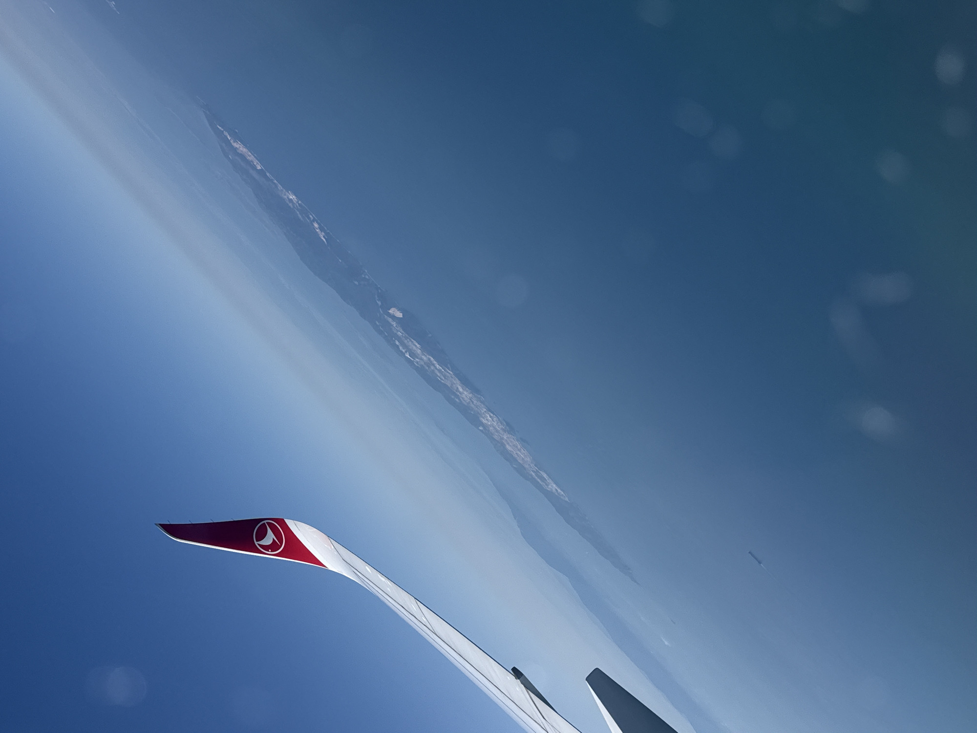 An aerial view from an airplane window shows a wingtip with a Turkish Airlines logo against a clear blue sky. Below the wing, the sea stretches out to a distant coastline, possibly a city, with visible lights and structures. A small ship is visible in the water. The image is tilted, suggesting the aircraft is banking.